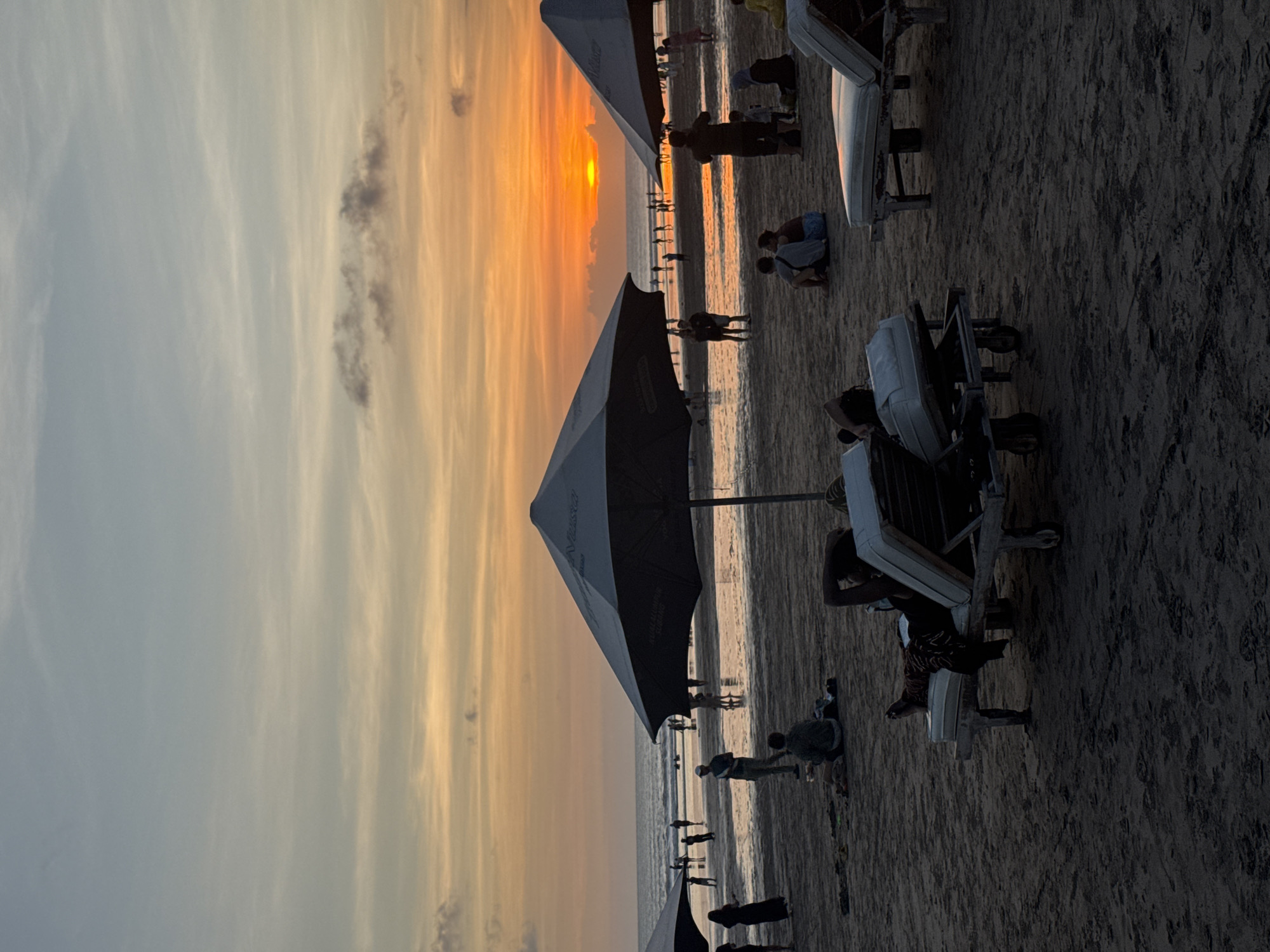 The image captures a sunset scene on a beach, potentially in Kuta, Indonesia, given the context. The sun is setting on the horizon, creating a vibrant orange and yellow sky. Silhouetted people are visible along the shoreline, some walking and others standing or sitting. Beach umbrellas and lounge chairs are positioned on the sand, with some individuals relaxing in them. The foreground features the sandy beach, while the background includes the sea and the colorful sky.
