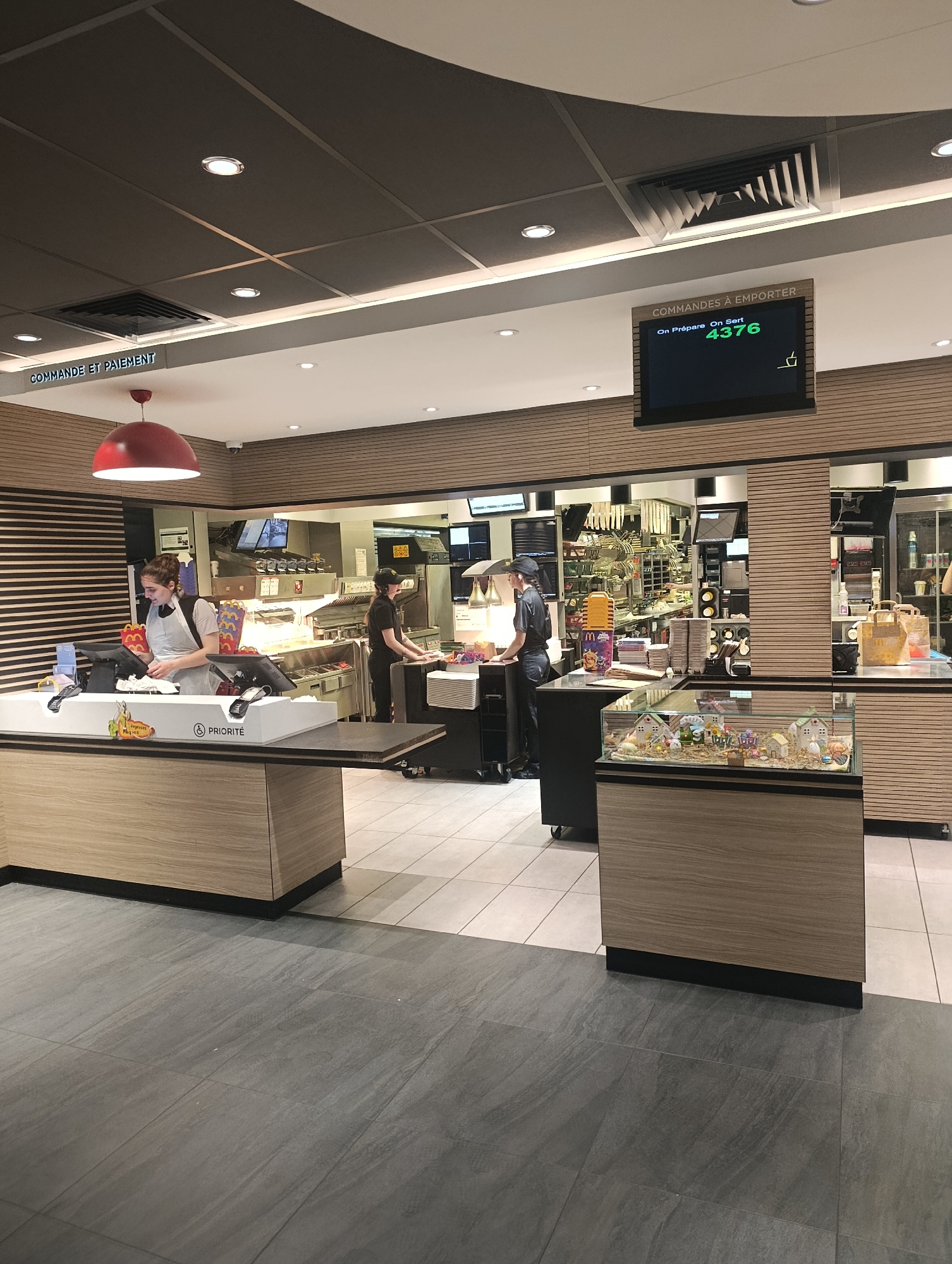 The interior of a McDonald's restaurant in Freyming-Merlebach, France, features a service area. A woman with her hair up is working behind the counter at a cashier station. Two employees, both wearing black uniforms, are filling orders near the back of the restaurant. Above the service area, a digital display reads "COMMANDES A EMPORTER" (Orders To Go) with the order number 4376 highlighted. The interior design includes wood-paneled walls, light-colored counters, and gray tile flooring. A display case with small figurines sits near the service counter.