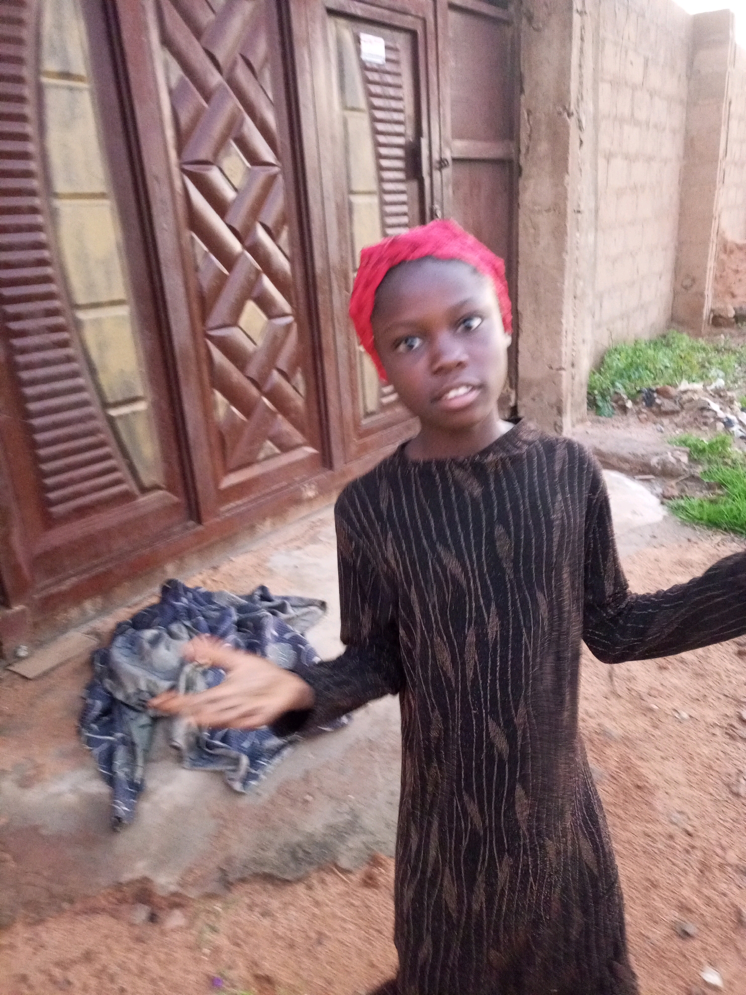 In Tempure, Nigeria, a young person wearing a red headscarf and a black patterned long-sleeved dress stands outside a building with large wooden doors. To their left, a blue and grey patterned cloth lies on the ground. The ground is composed of reddish-brown sand and some concrete. In the background, a textured brick wall is visible with some green vegetation and debris. The person is looking towards the camera with their right arm extended.
