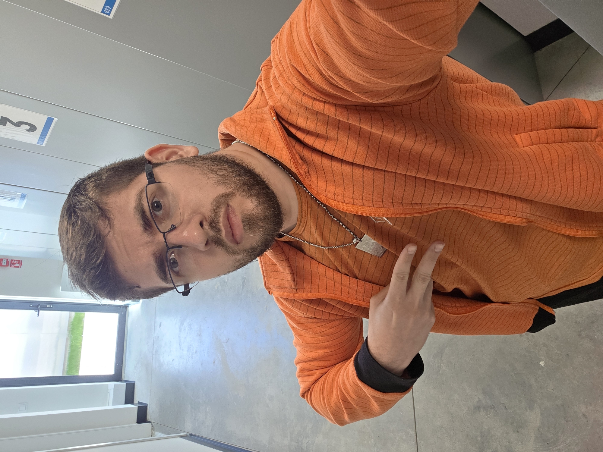 The image shows a man wearing glasses and an orange zip-up hoodie with black cuffs, standing indoors. He is wearing a silver chain necklace. He is making a peace sign with his right hand. A window with a view of green grass is visible behind him. The floor appears to be concrete. The location appears to be inside a building, possibly an office or public space, but it's impossible to confirm it's in Plovdiv, Bulgaria, without more information.