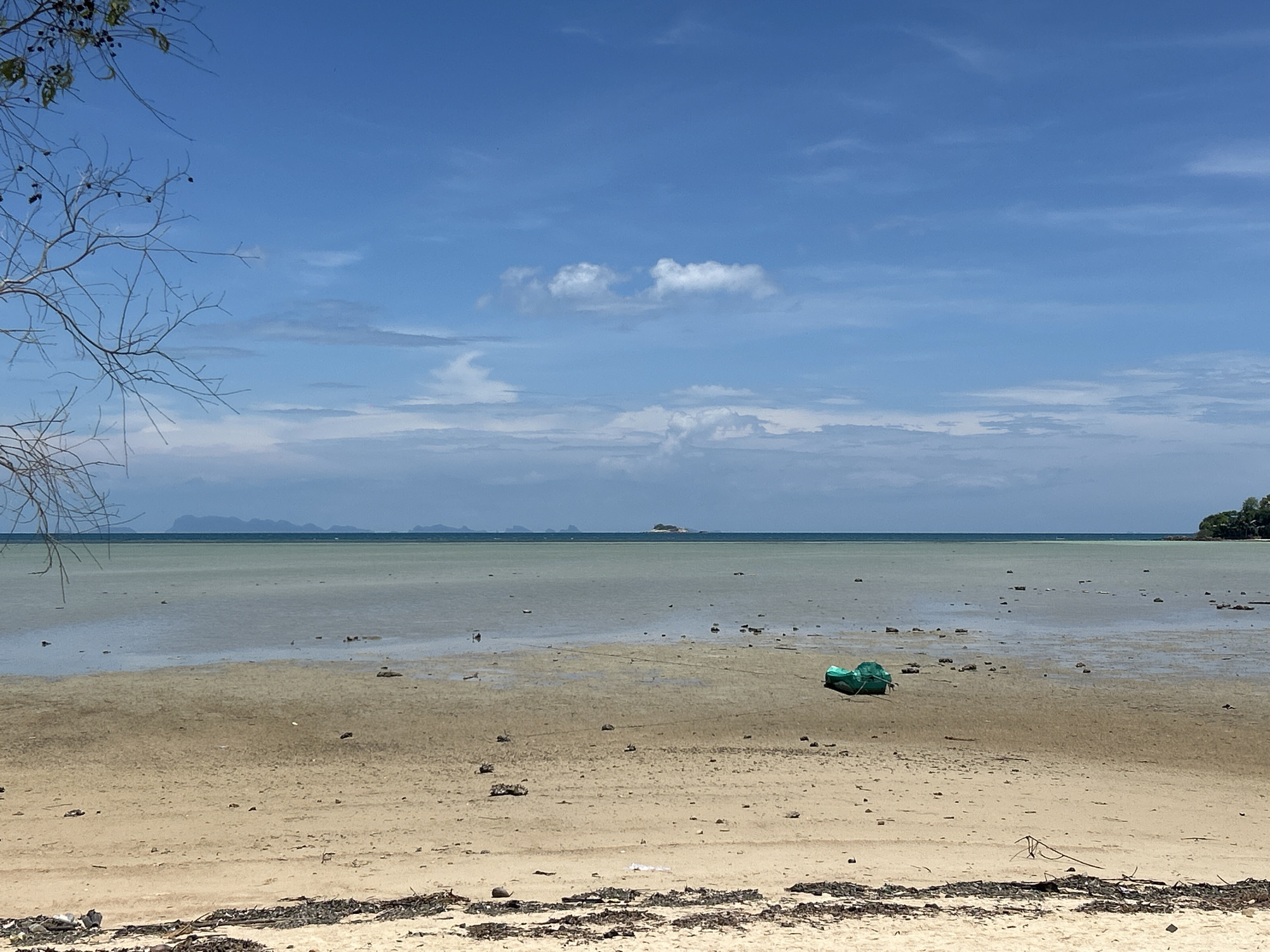 The image depicts a beach scene in ตำบลเกาะพะงัน, Thailand. The foreground features a sandy beach with scattered debris of wood. A low tide exposes a shallow, greenish expanse of water stretching towards the horizon, where distant landmasses and islands can be seen. A single, green boat is visible on the exposed sand. The sky is blue with scattered clouds. A bare tree branch is visible in the upper left corner.