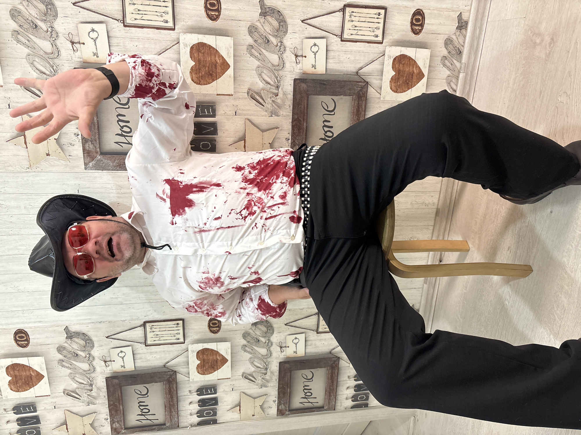 A middle-aged Caucasian male wearing a black cowboy hat, red sunglasses, and a white button-down shirt splattered with red, sits on a wooden stool in front of a white wooden wall decorated with various framed pictures, wooden hearts, and the word "hello" in metal letters. He is wearing black pants and a studded belt. His right hand is raised, palm facing forward, in a gesture that could be interpreted as a stop sign or greeting. The wall also features framed pictures with the word "Home" and other decorative elements. The floor is light-colored wood. No specific location within Krasnodar, Russia can be determined from the image.