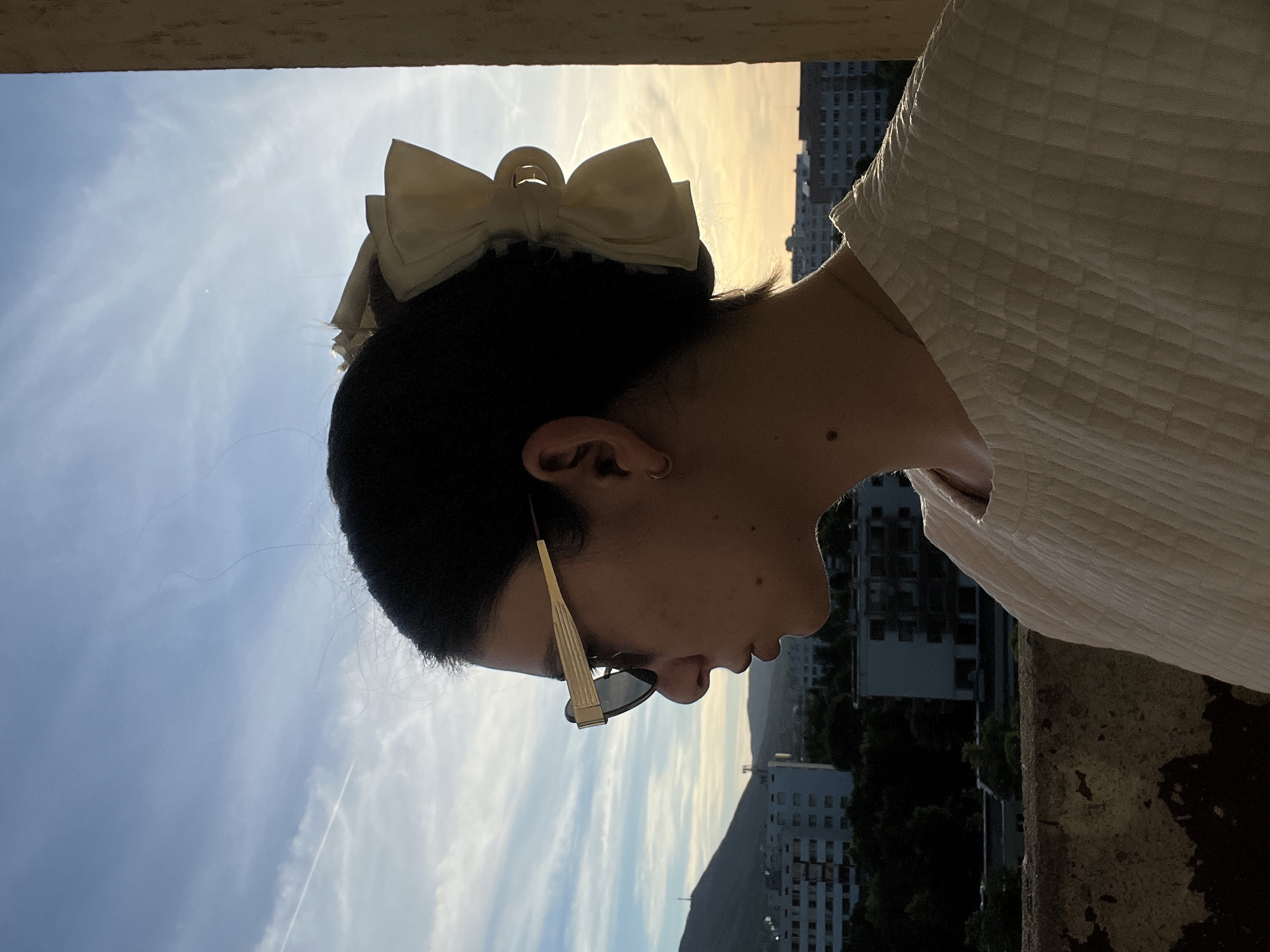 A young woman with dark hair styled in a bun with a large, light-colored bow is captured in a profile view against a dusky sky with scattered clouds. She is wearing large, stylish sunglasses with gold frames and a light-colored, textured top. She appears to be standing on a balcony or elevated platform, with a railing in the foreground. In the background, several multi-story buildings and a distant hill or mountain are visible, suggesting an urban or suburban setting. The lighting indicates either sunrise or sunset.