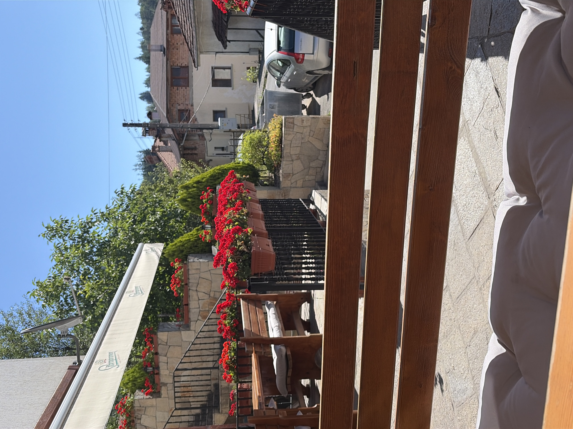 The image depicts an outdoor seating area, possibly a cafe or restaurant, in Smolyan, Bulgaria. In the foreground, a wooden bench with a light-colored cushion is visible, with a wooden railing in front of it. The middle ground features a rustic wooden table and bench set, adorned with multiple pots of vibrant red flowers. Behind the seating area, a metal railing separates it from a stone-tiled pathway. Beyond this, a silver car is parked near a large metal dumpster. The background reveals a street scene with several buildings, including houses with tiled roofs, and utility poles with electrical wires. Lush green trees and foliage are scattered throughout, and the sky is a clear blue. The overall impression is a sunny day in a European town.