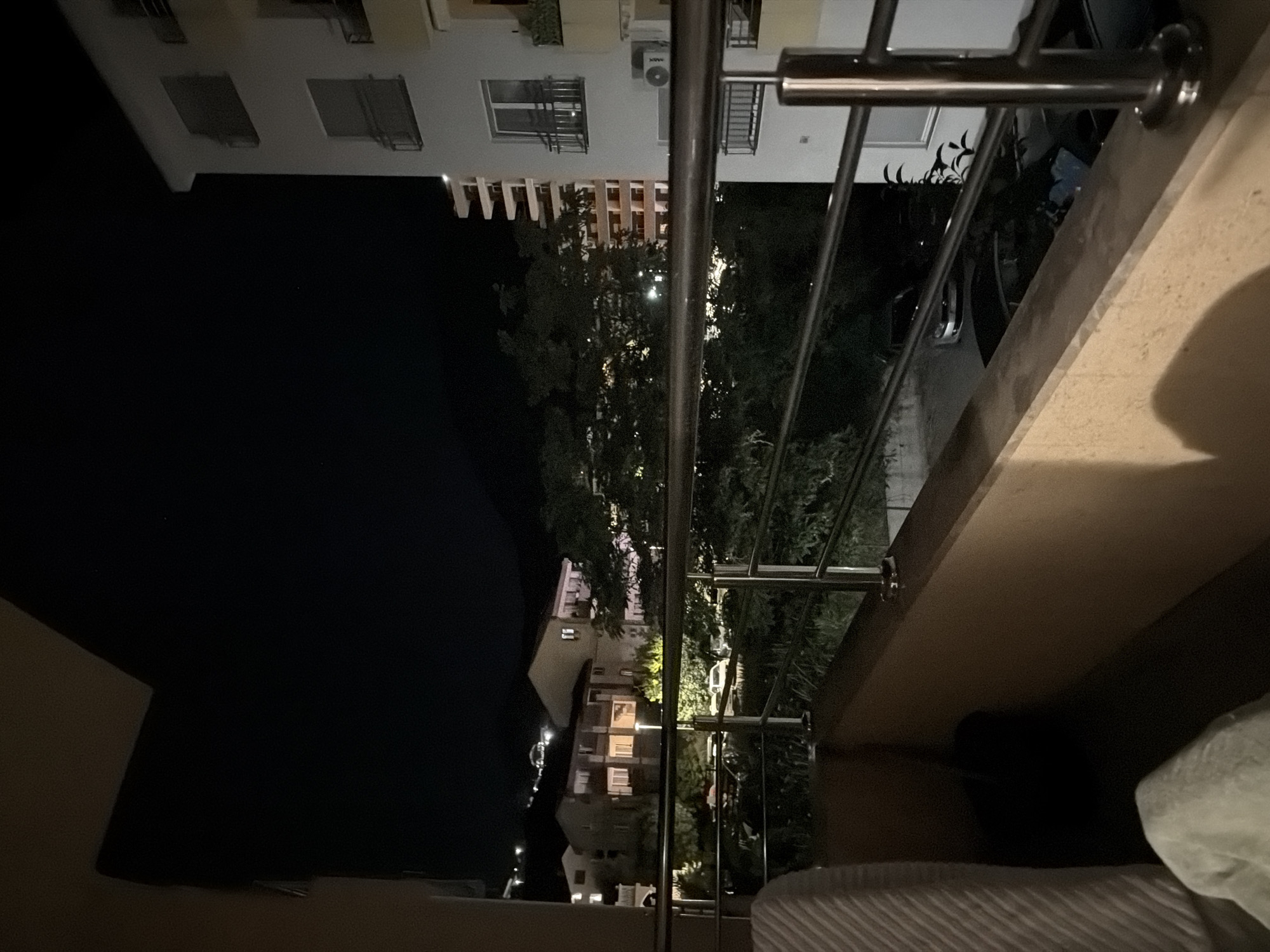 The image shows a night view from a balcony overlooking a residential area in Budva, Montenegro. In the foreground, a beige-colored balcony railing with stainless steel bars is visible. To the left, part of a light-colored, textured cushion or blanket is present. Beyond the railing, a street or parking area with several parked cars is illuminated by streetlights and interior lights of buildings. The cars appear to be dark-colored sedans.
Further in the background, there are several residential buildings of varying heights and architectural styles, some with visible lit windows. Trees and foliage are interspersed between the buildings, casting shadows and adding to the nocturnal atmosphere. In the distance, a dimly lit hillside or mountain can be seen. The sky is dark and clear, with a few faint stars visible. The overall impression is a quiet, urban night scene.