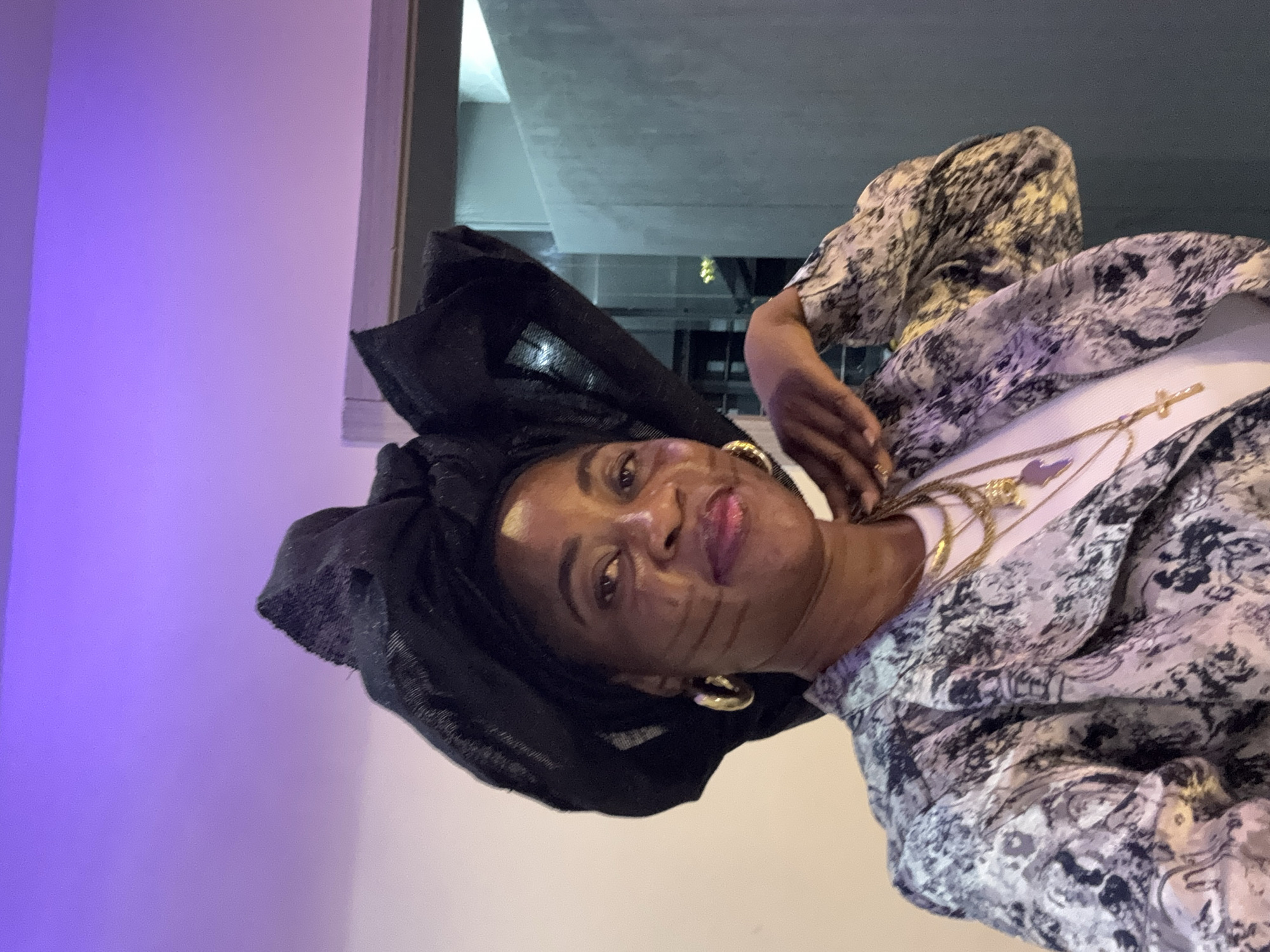 A medium shot shows a Black woman wearing a black headwrap, gold hoop earrings, a white t-shirt, and a patterned jacket. Her left hand rests on her chin. The background is a wall lit with purple light. The image appears to be taken indoors.
