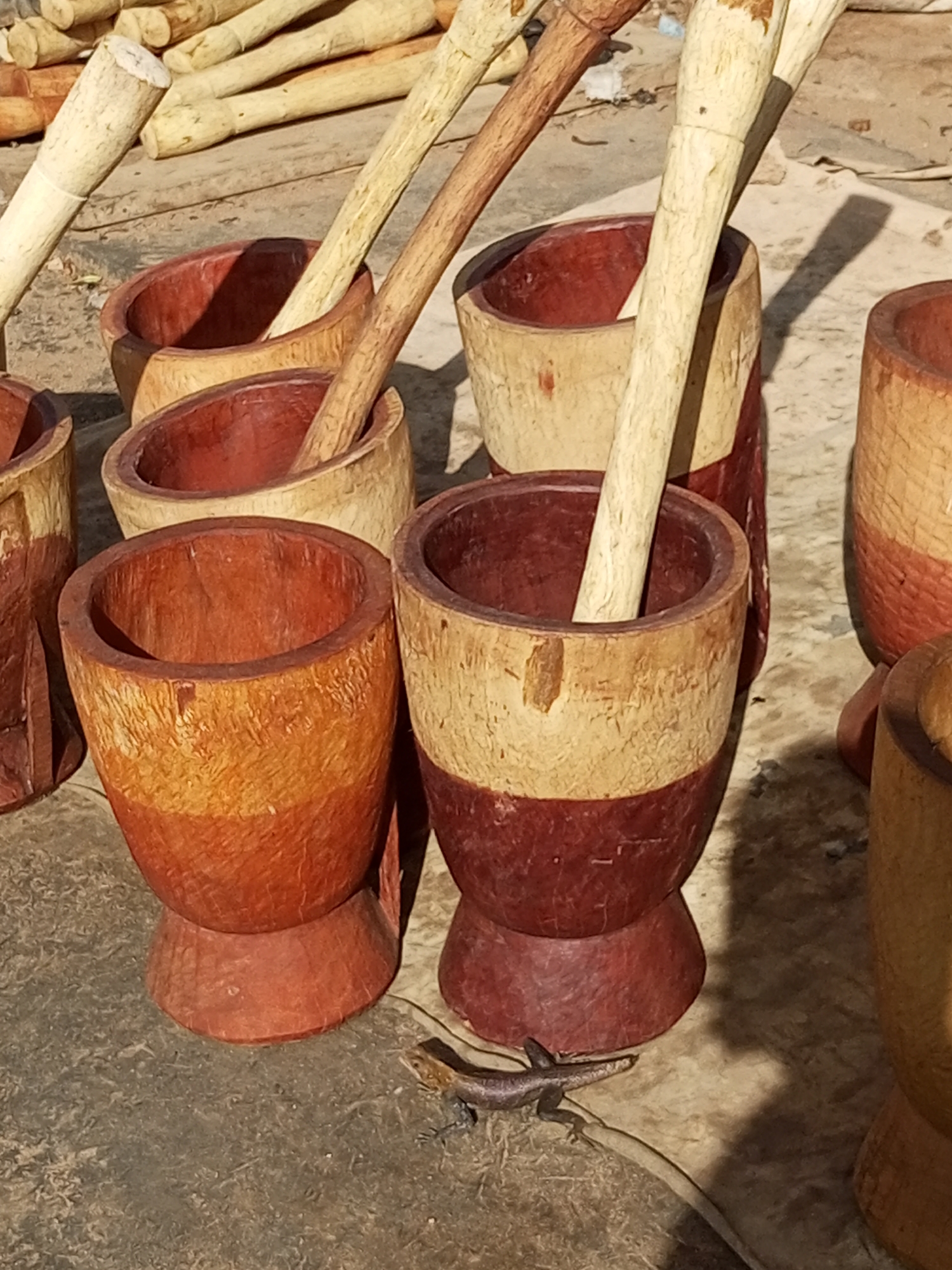 The image depicts a collection of handcrafted wooden mortars and pestles, likely for sale, on a dirt or stone surface in Arawa, Nigeria. Several mortars, varying in size and color, are visible, some with pestles resting inside or nearby. The mortars are primarily made of wood, with some showing a reddish-brown interior and a lighter, natural wood exterior, often banded with a darker reddish-brown at the base. The pestles are long, cylindrical wooden pieces. In the foreground, a small lizard with brown and grey markings is positioned on the ground between two of the mortars. In the background, more wooden objects, possibly unworked logs or parts of similar tools, are piled up. The lighting suggests it is daytime, with shadows cast by the objects indicating the sun's position.