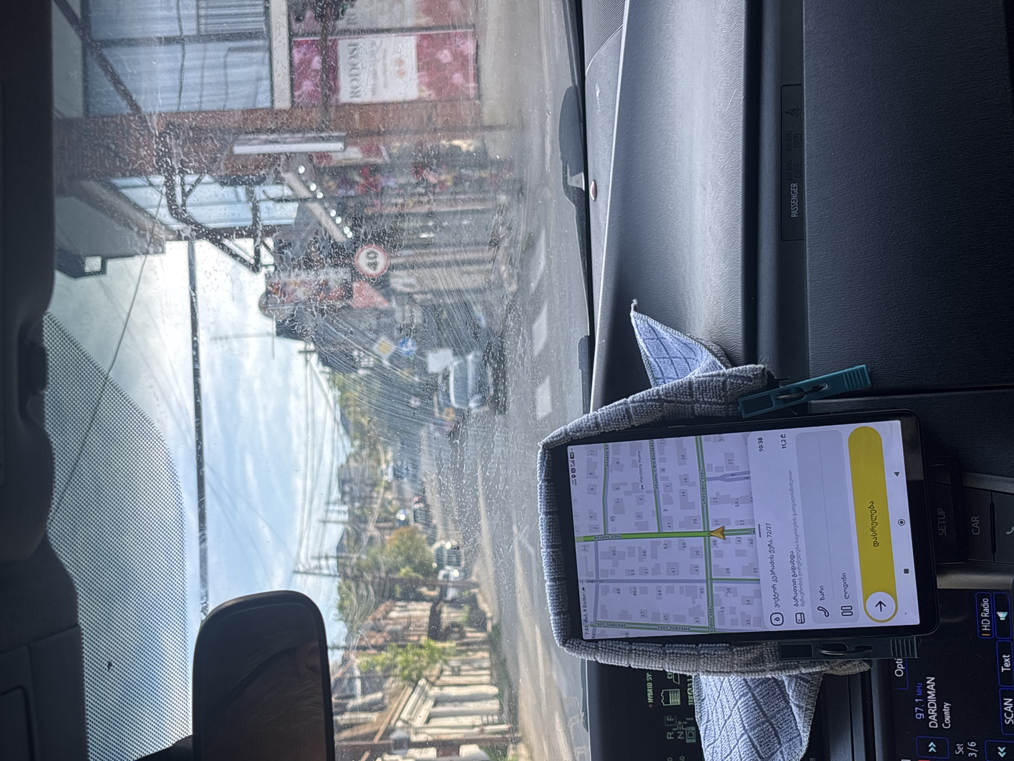The image shows the view from inside a car, presumably on a street in Tbilisi, Georgia. The windshield is somewhat dirty, obscuring the view. Through the windshield, a street can be seen with buildings and trees lining the sides. A "40" speed limit sign is also visible.
Mounted on the dashboard is a smartphone displaying a navigation app, seemingly guiding the driver to a destination on Viktor Kupradze Street 72/27. The navigation app is in Georgian. The smartphone is being held in place by a clothes pin and what appears to be a small towel.
The car's radio display is visible, set to 97.1 MHz, with the name "DARDIMAN" and "Country" displayed, and some of the dashboard. The passenger airbag indicator is also visible, set to "ON."