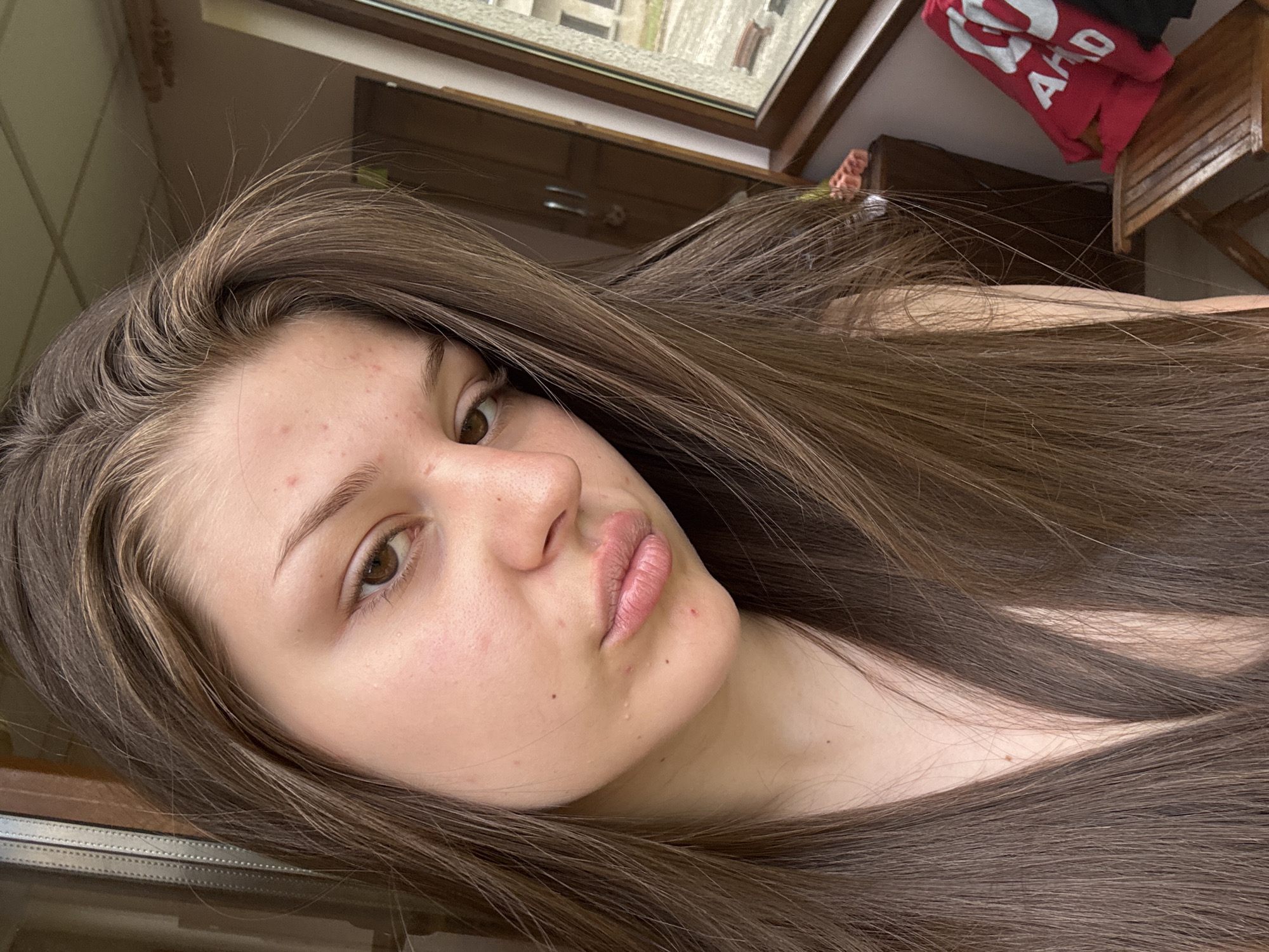 The media file is a close-up, overhead selfie of a young woman with long, brown hair. Her face is angled towards the camera, and her hair is spread around her. She has brown eyes, light skin with some blemishes, and is wearing light-colored makeup. Her lips are slightly parted, and she has a neutral expression. In the background, there is a window with a view of what appears to be a building, a wooden dresser or cabinet, and a wooden chair. A red shirt with white lettering is draped over the chair or dresser. The lighting suggests it is daytime, but the indoor setting makes it difficult to determine the exact time of day or weather. There is no discernible activity or event taking place, other than the woman taking a selfie.