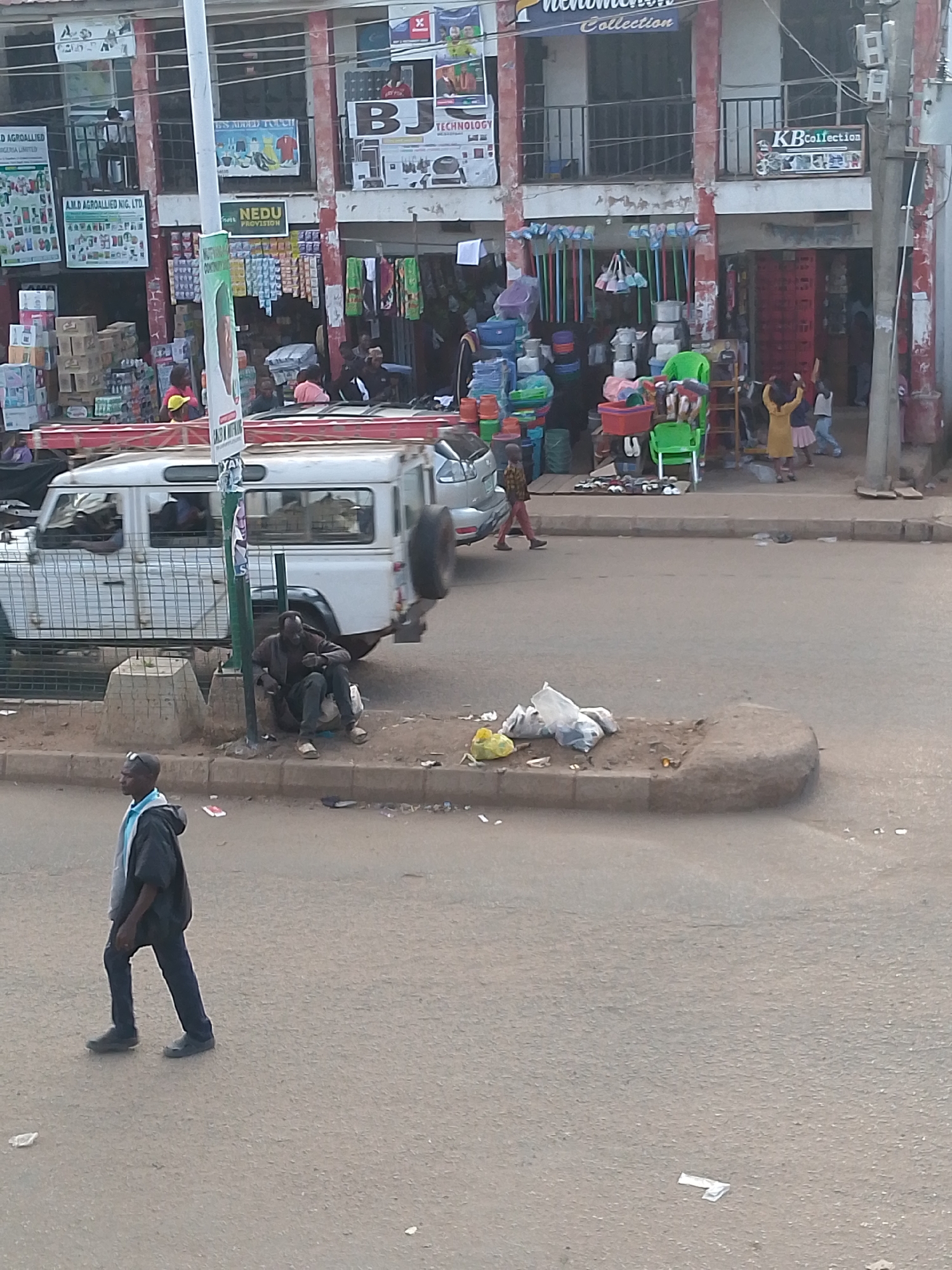 This outdoor image captures a lively street scene in Bukuru, Nigeria, characterized by bustling commercial activity. The setting features multi-story buildings with shops on the ground floor, distinguished by red structural elements and faded white walls.
In the mid-ground, a white Land Rover Defender-type vehicle, equipped with a red ladder on its roof rack and a spare tire mounted at the rear, is parked on the left side of the street. Adjacent to it, a man dressed in dark clothing sits on a concrete block beside a utility pole. In the foreground, another man wearing a dark jacket over a light blue shirt walks across the paved street from left to right. The street and sidewalk show signs of litter, including plastic bags and wrappers.
The shopfronts are filled with various goods, including brightly colored stacked plastic basins, buckets, brooms, and mops, indicating general merchandise stores. Cardboard boxes are piled high outside a shop on the left. Numerous individuals are present in and around the shops; two children, one in a yellow dress with raised arms, are visible near the rightmost shop, while others appear to be vendors or customers.
Visible text on the shop signs and banners includes "AMD AGROALLIED NIG, LTD.", "NEDU PROVISION", "BJ TECHNOLOGY", and "KB Collection". Partially visible text also includes "AGROALLIED NIGERIA LIMITED" and "phenomenon Collection". The scene is illuminated by daylight, suggesting a morning or afternoon setting, and portrays the dynamic atmosphere of an urban marketplace.