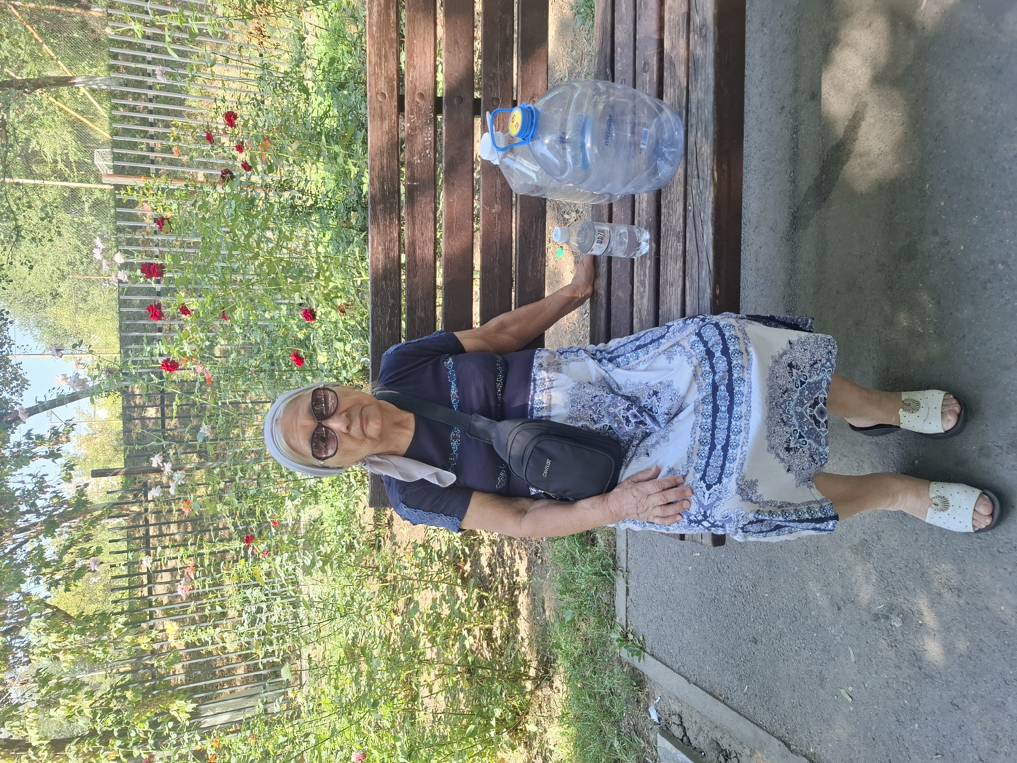 An elderly woman sits on a wooden park bench in Almaty, Kazakhstan, on a sunny day. She is wearing a dark blue t-shirt, a light blue and white patterned skirt, a light blue headscarf, and sunglasses. A black crossbody bag is slung over her shoulder. On the bench beside her are two large plastic bottles of water and one smaller bottle. Behind the bench is a dense green hedge with numerous red and pink roses, and a metal fence. The ground in front of the bench is paved with asphalt.