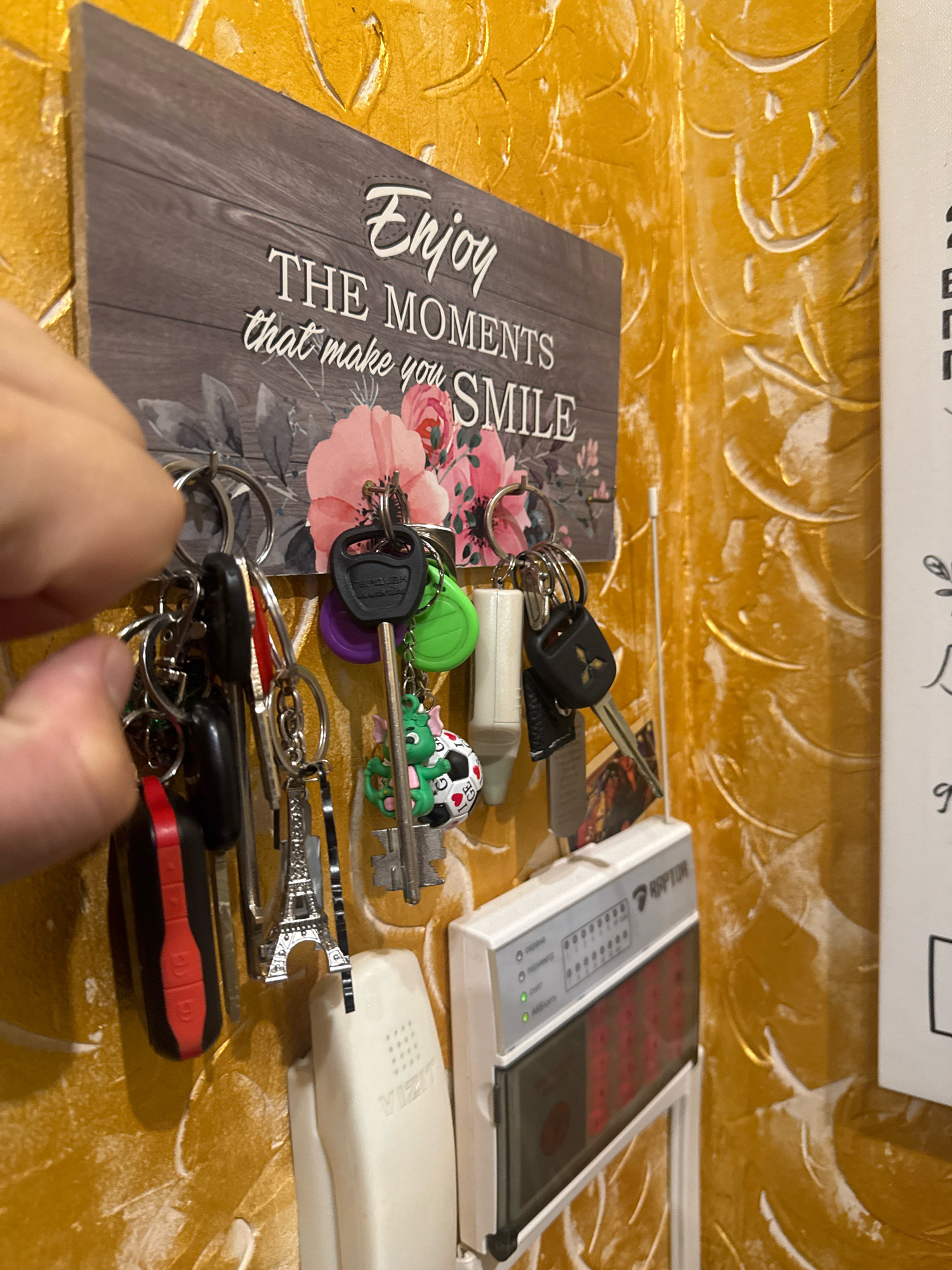 A hand is reaching for a set of keys hanging from a key holder on a textured yellow wall. The key holder is a wooden sign with the words "Enjoy THE MOMENTS that make you SMILE" and an image of pink flowers. There are several keys on the holder, including a key with a black Mitsubishi logo, a key with a green fob, a key with a purple fob, and a novelty key with a dragon charm. Below the key holder is a white intercom unit and a gray and white alarm panel. The wall has a rough, stucco-like texture and appears to be painted in a bright yellow color. The scene is indoors, likely in a residential hallway or entryway. The location is not specified as Almaty, Kazakhstan.