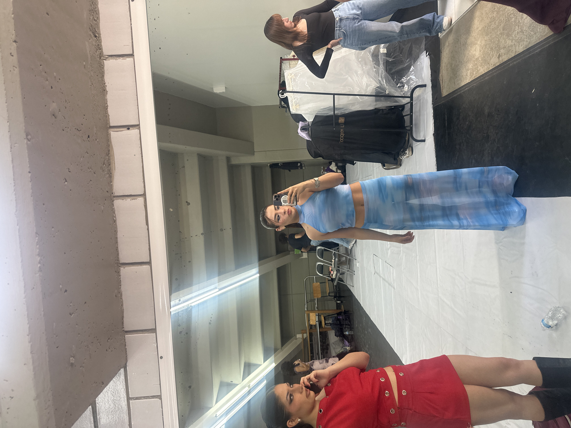 This image captures a mirror selfie taken by a young woman in an indoor setting, likely a studio or backstage area, in San Bernardino Tlaxcalancingo, Mexico. The original image is rotated 90 degrees counter-clockwise.
The main subject, reflected in the mirror, is a young woman with dark hair, wearing a light blue tie-dye crop top and a matching sheer, flowing skirt. She holds a smartphone, taking a photo of her reflection, and looks directly at the camera with a posed expression. Her midriff is visible.
To the upper right in the reflection, another woman with medium brown hair wears a black long-sleeved top and blue jeans, standing near a clothing rack covered in white plastic. Further back, another rack holds black garments, with partially visible text "coopni" on a bag.
In the lower part of the reflection, a third young woman with dark hair is seen lying or leaning back on a white tarp-covered floor. She wears a striking red two-piece outfit, featuring a cropped top and a mini skirt adorned with multiple silver grommets, paired with black boots. She looks towards the camera with a relaxed expression. A plastic water bottle, with "Cristal" partially visible on its label, rests on the white tarp near her.
The environment features concrete walls on the left, next to a section of white rectangular tiles bordering the large mirror. The floor is predominantly covered with white tarps, suggesting a setup for an event, photoshoot, or performance. Overhead fluorescent lighting fixtures are reflected in the mirror. In the deeper background, additional people, chairs, and tables are faintly discernible, indicating a busy, collaborative atmosphere. The overall scene conveys a behind-the-scenes moment.