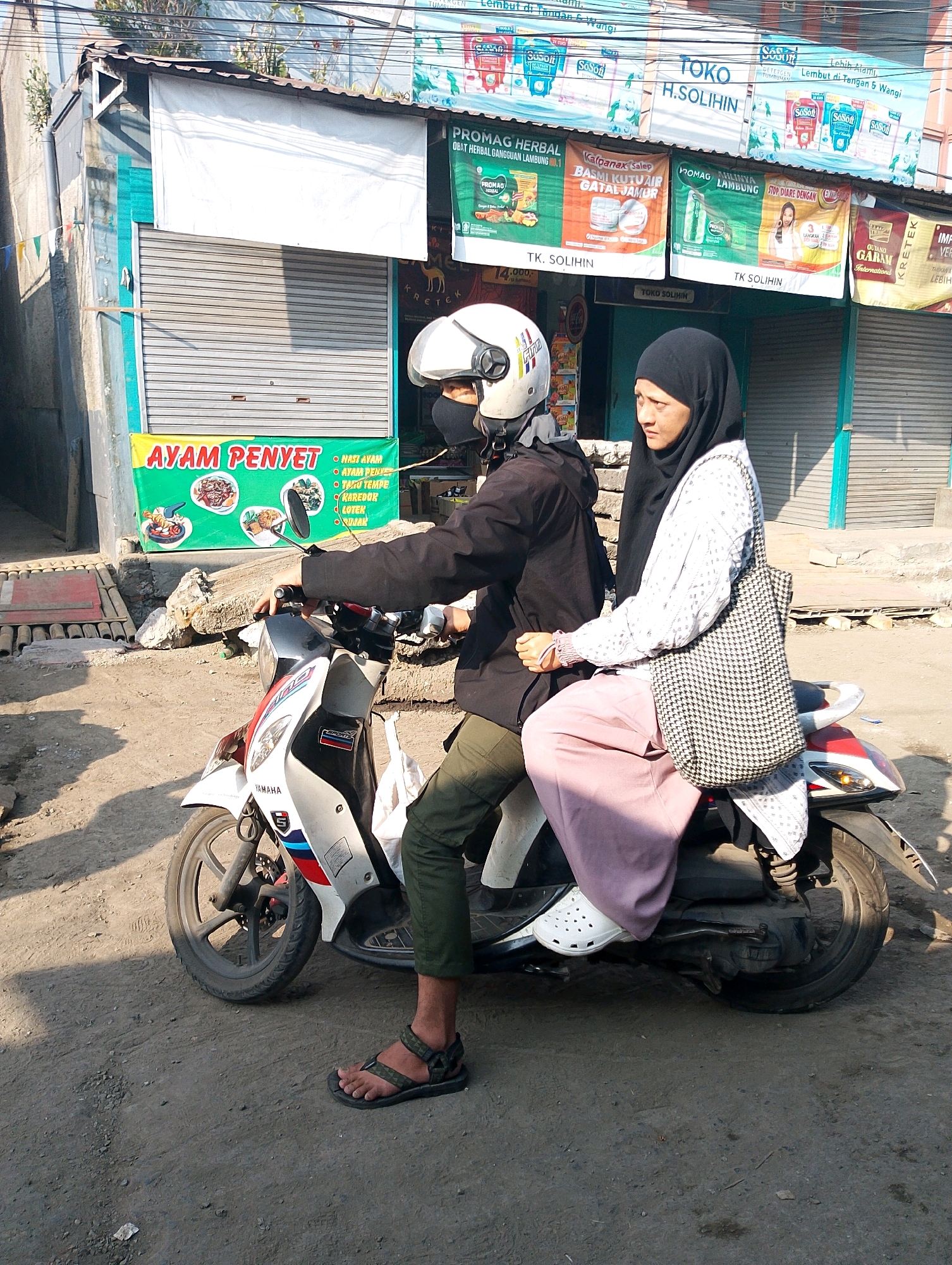 In Lembang, Indonesia, two individuals ride a scooter on a dirt road. The person in front, wearing a black jacket and white helmet, operates the vehicle. The passenger behind, a woman in a black hijab and a white patterned blouse with a black and white checkered bag, sits behind the driver. She wears a pink skirt and white Croc-style shoes. In the background, a storefront displays various advertisements, including one for "AYAM PENYET" (Smashed Chicken). The scene appears to be a typical street view in a commercial area.