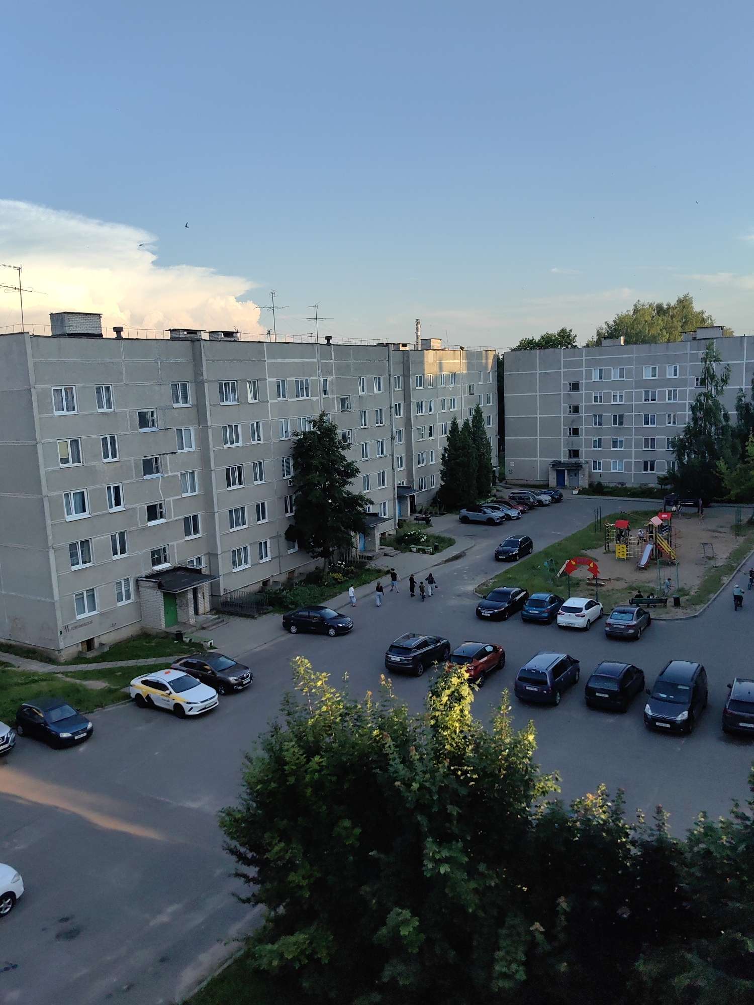 The image shows an aerial view of a residential area with two apartment buildings and a playground in Fanipal, Belarus. The buildings are typical Soviet-era panel buildings, with multiple floors and rows of windows. The apartment building on the left is taller than the one on the right. In front of the buildings, there is a paved area with many parked cars. Some people are visible walking around the buildings and near the playground. The playground has slides, swings, and a sandbox, and appears to be in use with some children and adults present. The sky is partly cloudy with a light blue hue, and the sun appears to be setting, casting long shadows. Trees and greenery are scattered around the buildings and playground, indicating a suburban or park-like setting. The overall scene depicts a typical evening in a residential neighborhood.