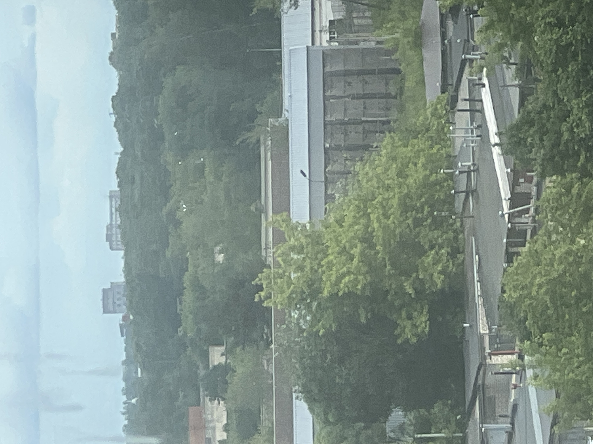 An aerial view captures a dense urban landscape dominated by greenery and industrial structures. In the background, two large, multi-story buildings with flat roofs are visible, possibly residential or commercial complexes. The middle ground features a long, low-rise industrial building with a corrugated metal roof and a series of vertical concrete elements. This building is flanked by mature trees, some of which are lush and green, while others appear sparser. In the foreground, a cluster of smaller, flat-roofed structures with visible ventilation pipes and what appear to be satellite dishes or other rooftop equipment are present. These structures are interspersed with more trees and foliage. The overall impression is one of an urban area that has integrated significant green spaces within its industrial and residential zones. The sky is overcast with a muted, hazy quality, suggesting a cloudy or humid day.
