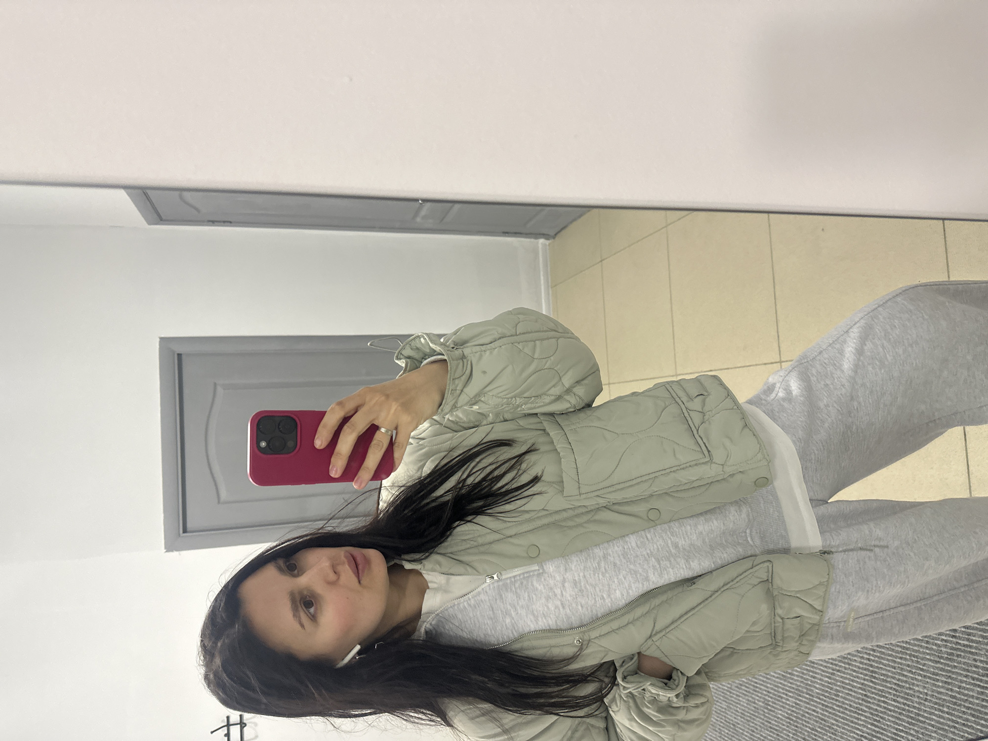 A woman is taking a selfie in what appears to be a dressing room or bathroom. She is holding a red phone and looking at the camera. She is wearing grey sweatpants, a white top, and a light green quilted jacket. The background includes a door, a tiled floor, and a mirror reflecting these details. Without additional information or reference points, it is not possible to confirm the specific location as Chaikovsky, Russia, or any other location.