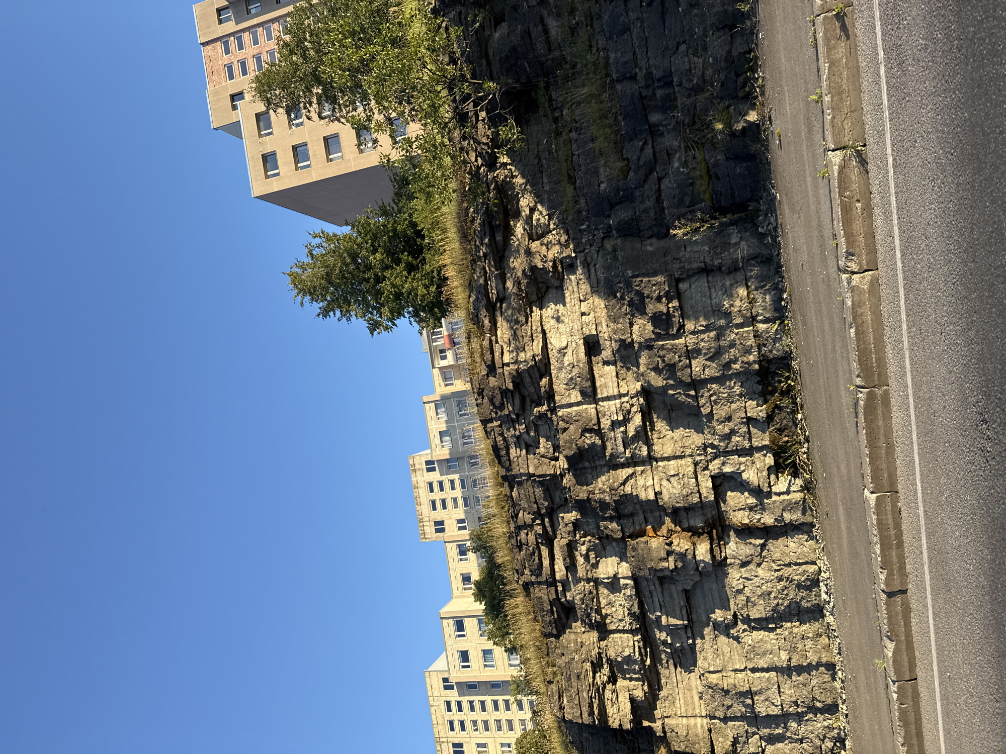 The image shows a view of apartment buildings on a sunny day in Tallinn, Estonia. In the foreground, there is a paved road with a concrete curb, and a rocky cliff face rising above it. The cliff face is layered and shows signs of erosion. Behind the cliff, several multi-story apartment buildings are visible, with a clear blue sky overhead. Trees and dry grass grow on top of the cliff and around the buildings. The scene appears to be located in a residential area, possibly in the Lasnamäe district, which is known for its Soviet-era apartment blocks and distinctive landscape features. No people or vehicles are present in the image.