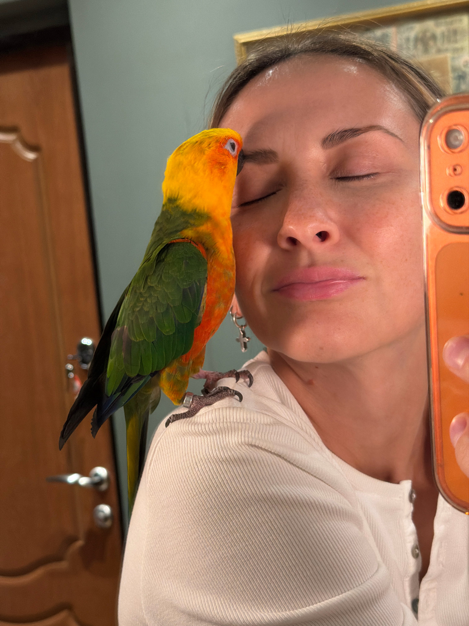 A person is taking a selfie with a sun conure parrot perched on their shoulder. The person's eyes are closed, and they are smiling slightly. The parrot is facing the camera, with its head tilted slightly. The background is a room with a wooden door and a framed picture on the wall. The image appears to be taken indoors. There is no information to indicate this scene is located in Moscow, Russia.