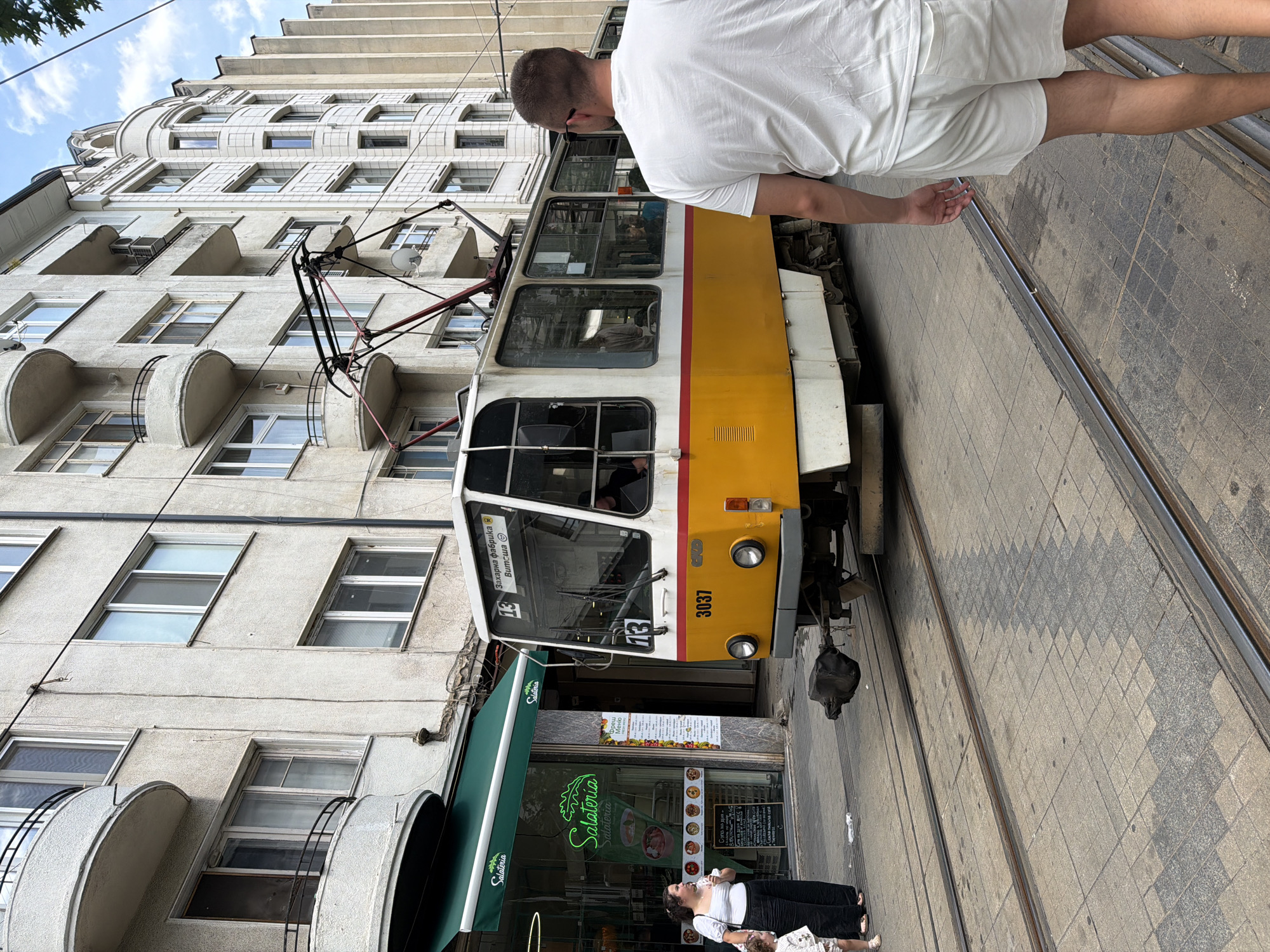 A man in a white t-shirt and white shorts is walking away from the camera on a city sidewalk in Sofia, Bulgaria. He is walking parallel to a yellow and white tram that is stopped at what appears to be a tram stop. The tram has the number 13 displayed prominently on the front and sides. To the left of the tram is a store with a green awning and a sign that reads "Salateria." A woman and a young child are standing near the store entrance. The buildings in the background are made of concrete and have multiple windows and balconies. The sky is blue with some clouds. The ground is paved with cobblestones and has tram tracks running through it.