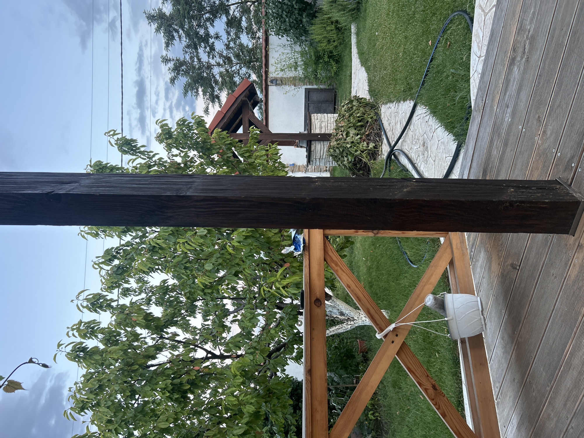 The image shows a view of a backyard in the village of Voyvodino, Bulgaria, from a wooden deck. In the foreground, a dark wooden pillar is visible on the right, and a wooden railing with a diagonal cross brace is on the left. A white planter pot hangs from the railing by a white string. Beyond the railing, a green lawn stretches to a paved pathway. To the left of the pathway is a well with a wooden roof structure and stone construction. A pile of green cuttings is next to the well. In the background, a white wall with a dark metal door is visible, with vegetation and trees beyond it. The sky is overcast with a light blue hue.