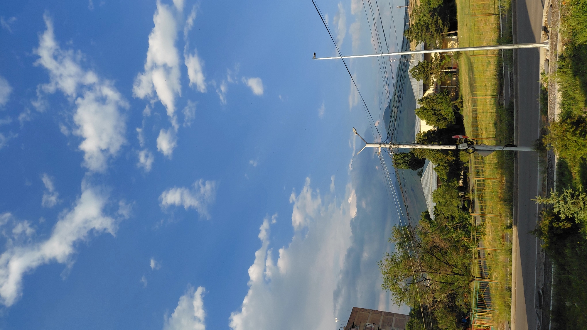 The image depicts a street view in Abovyan, Armenia, under a bright blue sky with scattered white clouds. In the foreground, a paved road runs horizontally across the bottom of the frame, bordered by green grass and a small evergreen sapling. Behind the road, a yellow metal fence separates the street from a grassy area with scattered trees and some buildings. A streetlamp with multiple lights is visible on the right side, and another streetlamp is partially visible on the left. Power lines crisscross the scene. In the background, a large building with a light-colored roof is visible, surrounded by trees. Beyond the buildings, a mountain range is faintly visible under the clouds. The overall impression is a quiet, suburban or semi-urban environment in Abovyan, Armenia.