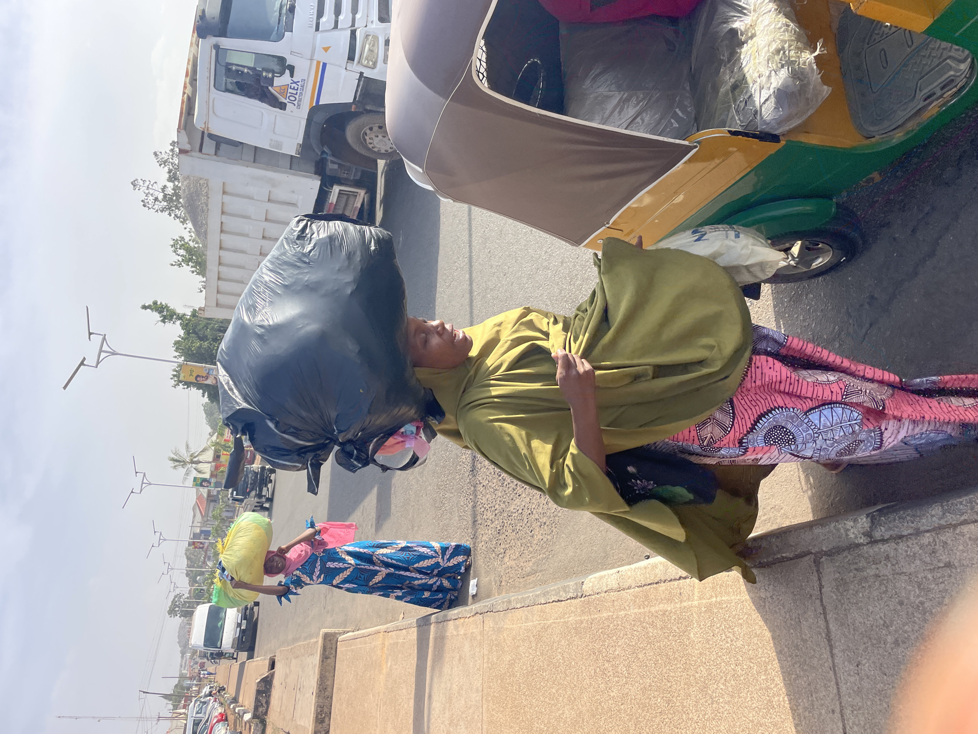 In Jos, Nigeria, a woman carrying a large black plastic bag on her head stands beside a green and yellow auto rickshaw on a paved street. She is wearing a green garment and a long, pink patterned skirt. Another woman walks in the background, also carrying a yellow bag on her head and wearing a blue patterned dress. A white dump truck is parked nearby with the logo "JOLEX Constructor CO. LTD." on the side. The environment suggests a busy urban area.