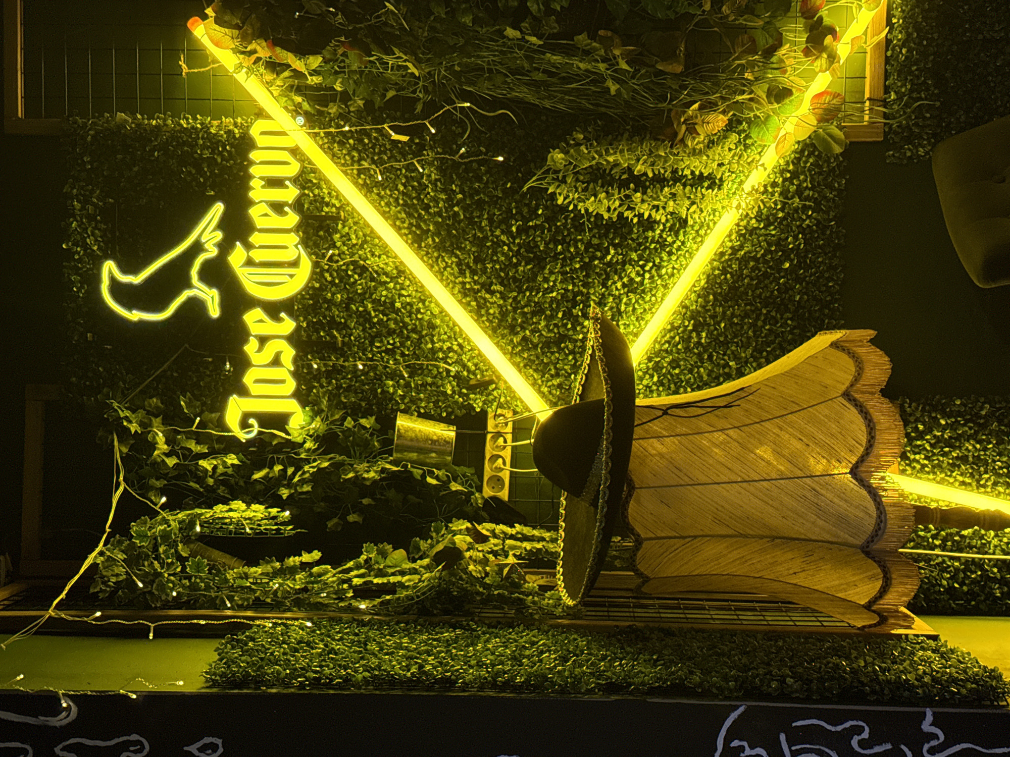 The image displays an interior or covered outdoor space, likely a bar or restaurant, in the city of Unknown, Unknown. The dominant feature is a wall covered extensively with artificial green foliage, resembling boxwood hedges and hanging vines. Various lighting elements are integrated into this wall. A prominent yellow neon sign reads "Jose Cuervo" in a stylized font, with a yellow neon silhouette of a bird (likely a crow) positioned directly above the text. Two bright yellow neon light bars are arranged diagonally across the foliage wall, intersecting near the center-right. String lights with small white bulbs are strung across the foliage and hanging vines. In the lower-left foreground, a vintage-style standing lamp with a yellowish-brown fabric shade, featuring a fringed bottom edge, is partially visible. A black sombrero with decorative trim is placed on the lamp's stand, below the shade. To the right of the sombrero, positioned against the foliage wall, is a dark panel containing multiple electrical outlets (at least three visible), with a shiny metal bucket or container resting on top of an associated dark structure. Wires are connected to the outlets. The far-left edge of the image shows a dark wall, possibly a blackboard, adorned with white chalk drawings depicting abstract shapes and outlines. The scene is illuminated primarily by the neon and string lights, suggesting an evening or nighttime setting.