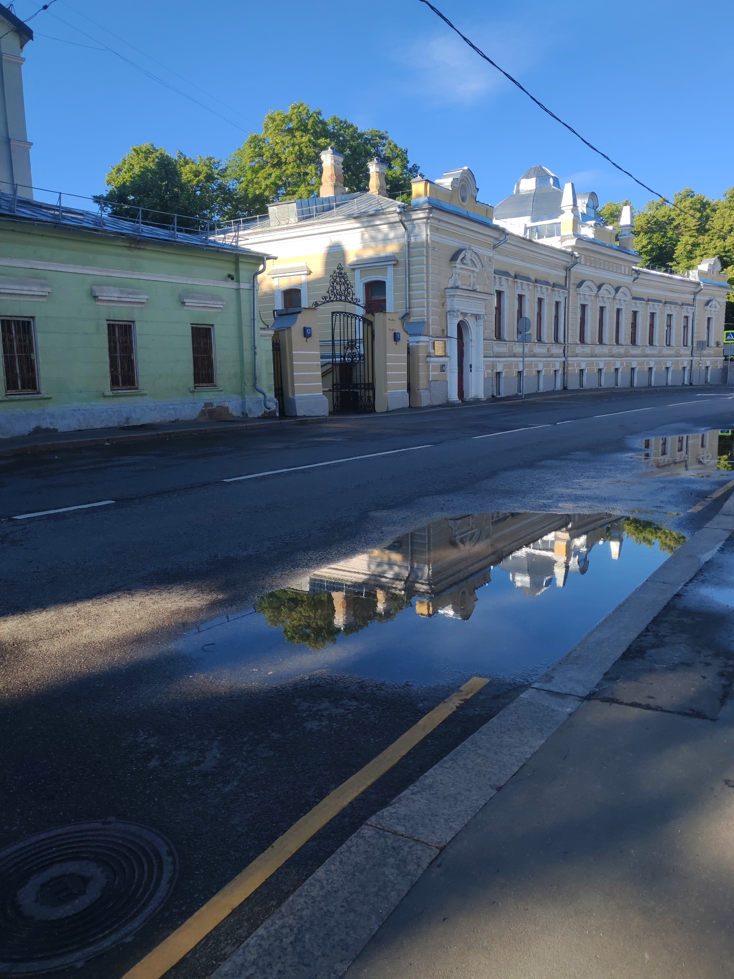 The image captures a street scene in Moscow, Russia, on a clear day with a bright blue sky. The focus is on the facade of a building with a light yellow color and intricate architectural details, featuring multiple windows with red frames and decorative elements above them. Adjacent to this building is a shorter, light green structure with barred windows. The buildings are situated along a wet asphalt road, indicated by a large puddle reflecting the buildings and trees. A sidewalk runs along the edge of the road, separated by a curb. A circular manhole cover is visible on the asphalt. In the background, lush green trees are visible, and power lines traverse the sky. The overall impression is one of an urban environment with historical architecture, suggesting a well-maintained street after a recent rain.