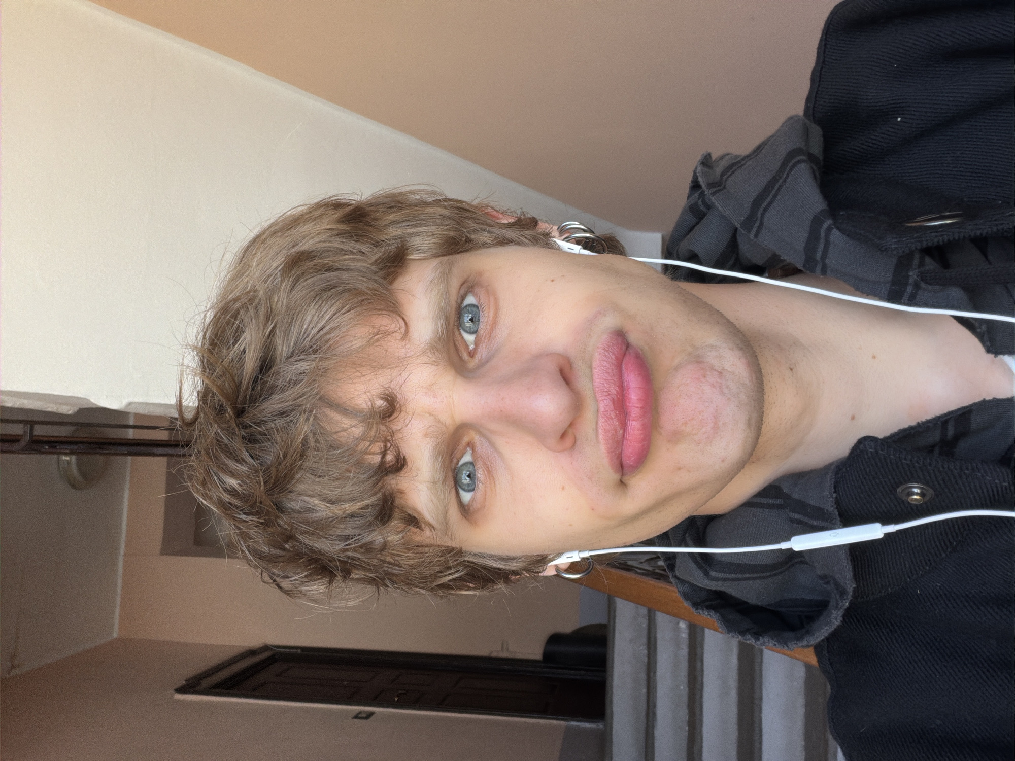 A Caucasian male with light brown, wavy hair and blue eyes is shown in a close-up, rotated at an angle. He is wearing white earphones and a black jacket with a grey plaid collar. The background consists of an interior space with beige walls and a glimpse of a staircase. Based on the architecture and the interior finishes, the location is potentially inside a residential or commercial building in Milan, Italy. No specific street or landmark is identifiable, and the description is inferred from the limited environmental context. The subject's expression is neutral.