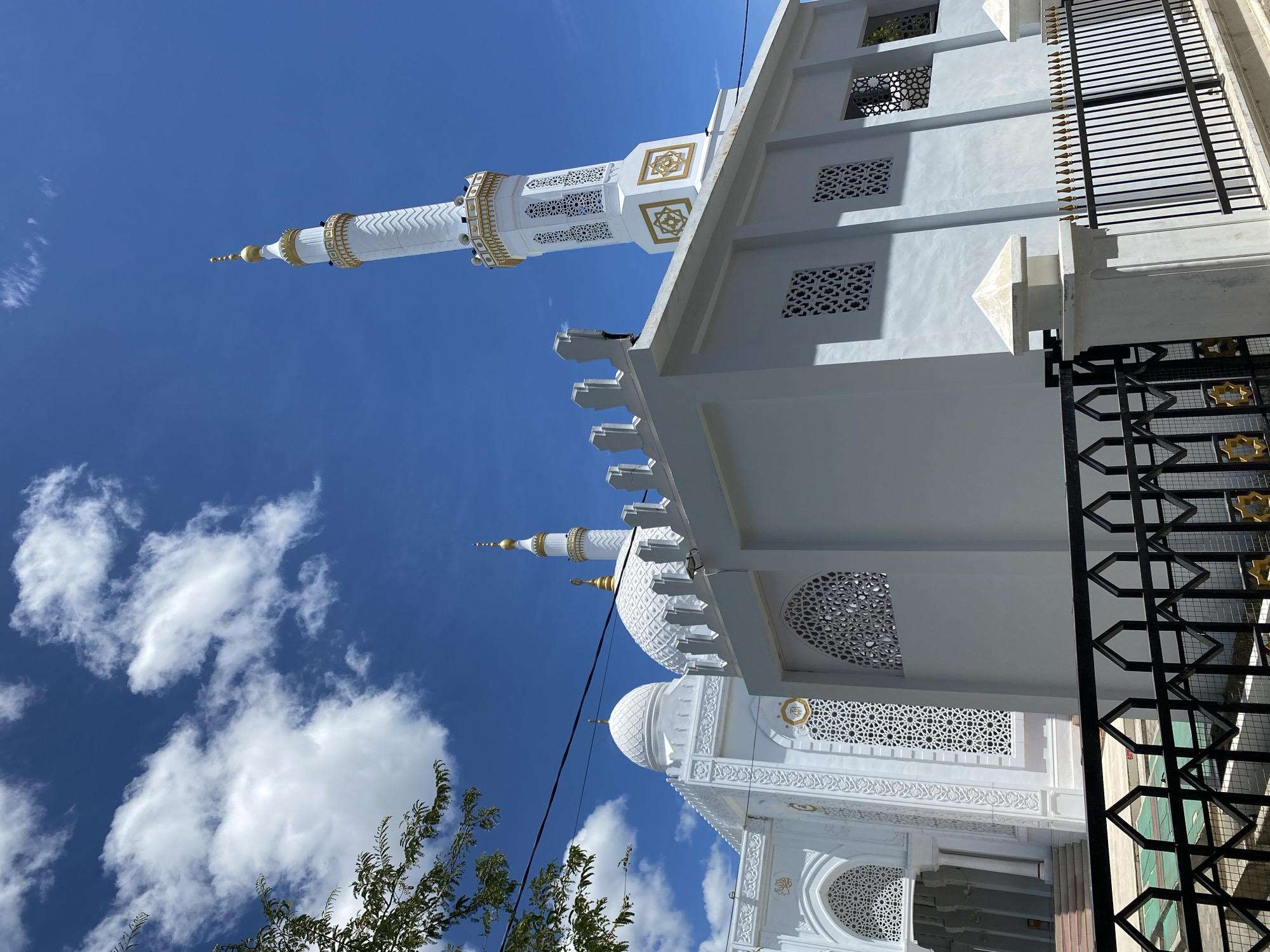 This is a low-angle, eye-level shot of a white mosque with gold accents, located in the city of Bayu, Indonesia. The mosque features multiple domes and a tall minaret, all of which are adorned with intricate patterns and decorative elements in white and gold. The building has a modern architectural style with sharp lines and geometric designs, particularly evident in the latticed windows and the minaret's structure.
In the foreground, a black metal fence with decorative gold elements runs across the bottom of the frame, separating the viewer from the mosque. Behind the fence, a white wall and a small white spire are visible. The mosque's facade is predominantly white, with ornate carvings and circular gold emblems.
The sky above is a vibrant blue with scattered white, puffy clouds. A few branches and leaves from a tree are visible on the left side of the image, creating a slight framing effect. The lighting suggests it is daytime, with clear sunlight illuminating the scene. There are no people visible in the image. The overall impression is one of a clean, well-maintained, and aesthetically striking religious building.