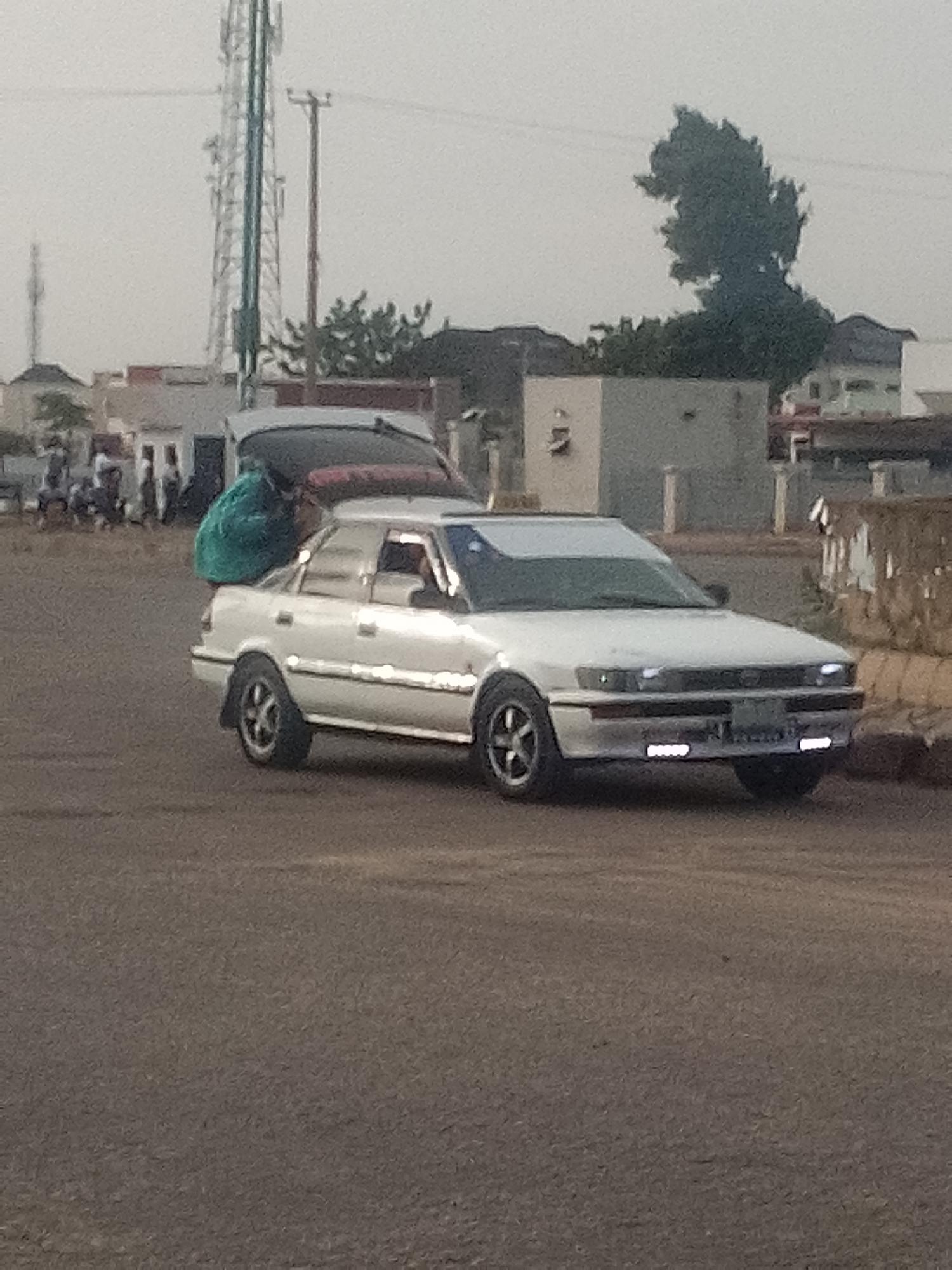 In Pantame, Nigeria, a white sedan, possibly a Toyota Corolla, is parked on a paved road, with its trunk open. A person wearing a green garment is placing a large green bundle into the open trunk. In the background, several other individuals are visible, appearing to be interacting or observing. The environment includes a cell tower, utility poles, and various buildings and trees, suggesting an urban or semi-urban setting. The sky is overcast, indicating a cloudy or hazy day. The scene appears to be a daytime event, likely involving the loading or unloading of goods.