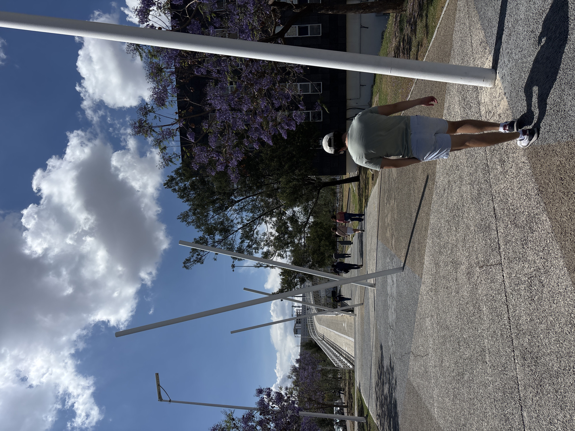 The image shows a man standing on a textured pavement, likely concrete, adjacent to a walkway lined with white metal poles. The poles create a geometric pattern against the sky, which is blue with puffy white clouds. Jacaranda trees with purple flowers are also visible. In the background, there is a bridge structure. A dark-colored building with multiple windows is situated behind the jacaranda trees. Several other people are walking on the walkway. The location could be Parque Lineal, Puebla, Mexico, where similar structures and trees can be found.