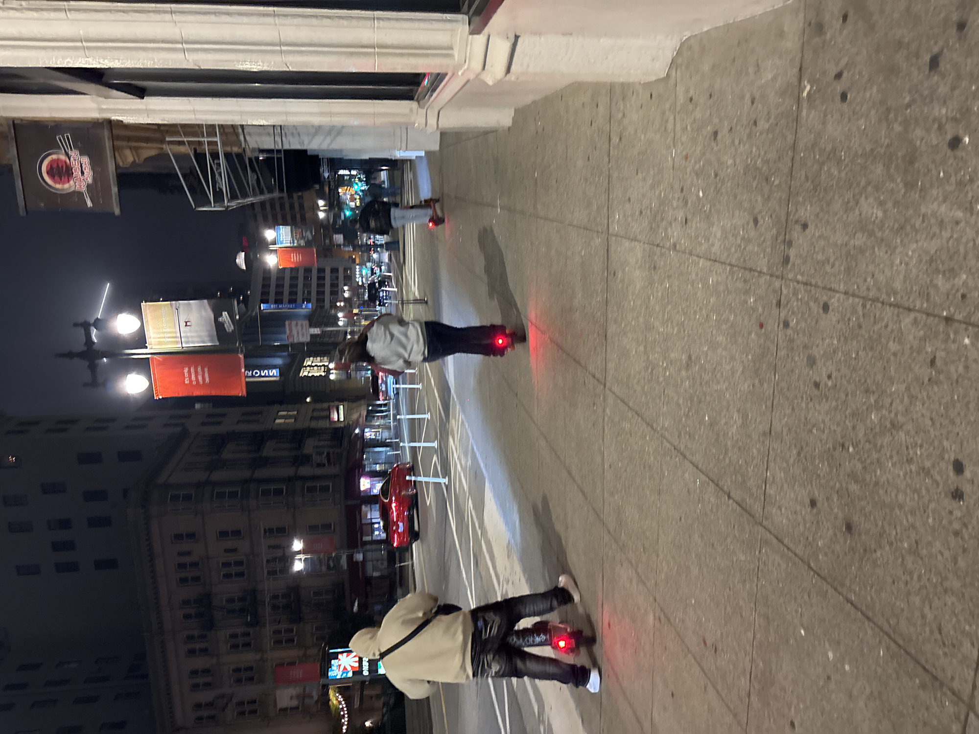 The image shows a nighttime street scene in San Francisco, California. In the foreground, a person wearing a beige hoodie and dark jeans rides an electric scooter with a red taillight. The person is facing away from the camera, looking down the sidewalk. Behind them, another person on an electric scooter with a red taillight is also moving away from the camera. Further down the street, a third person on a scooter can be seen. The sidewalk is paved with square tiles. Across the street, there are buildings, streetlights, and traffic. One building has a sign that appears to be for "RAISEN SUSHI". A red car is visible on the street. The overall atmosphere is urban and illuminated by streetlights and building lights.