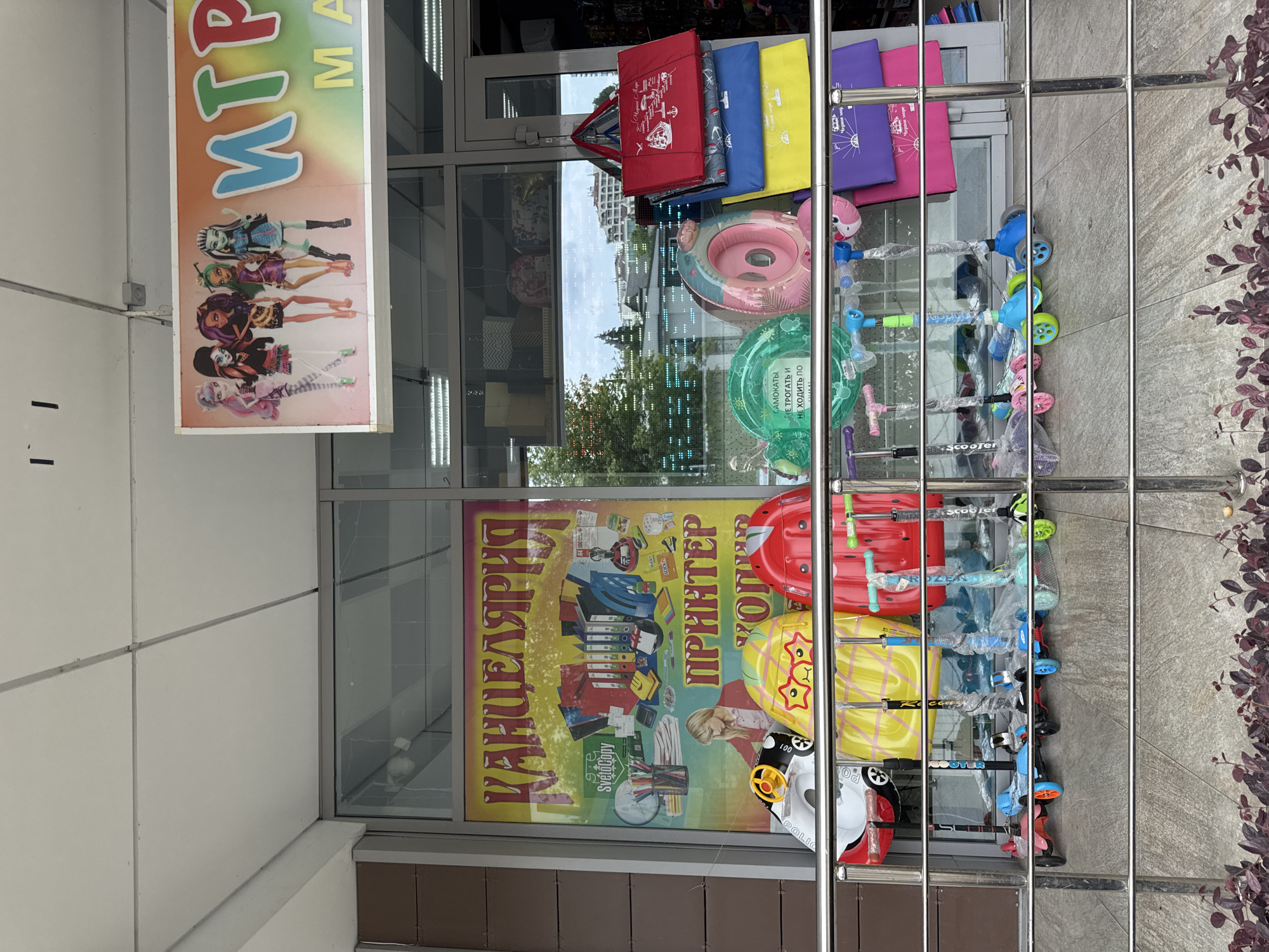 This image captures a storefront display, likely in Sochi, Russia, featuring a variety of children's toys and back-to-school supplies. In the foreground, a metal railing separates the viewer from the display. Behind the railing, several children's scooters are neatly arranged, some still in plastic packaging. Above the scooters, a colorful display showcases inflatable pool rings, one shaped like a snail and another pink.
Dominating the center of the image is a large poster advertising "КАНЦЕЛЯРІЯ" (Stationery) and "ПРИНТЕР" (Printer), with graphics depicting office supplies and a woman using a computer. To the right, a bright, rainbow-colored sign proclaims "ИГР" (likely part of a word for "toys" or "games"). On the far right, several colorful tote bags are stacked, red, blue, yellow, and purple, with nautical-themed graphics on some.
In the background, through the glass storefront, shelves with various items are visible, along with a glimpse of buildings and trees, suggesting an urban environment. The overall impression is a retail setting geared towards families and back-to-school shopping.