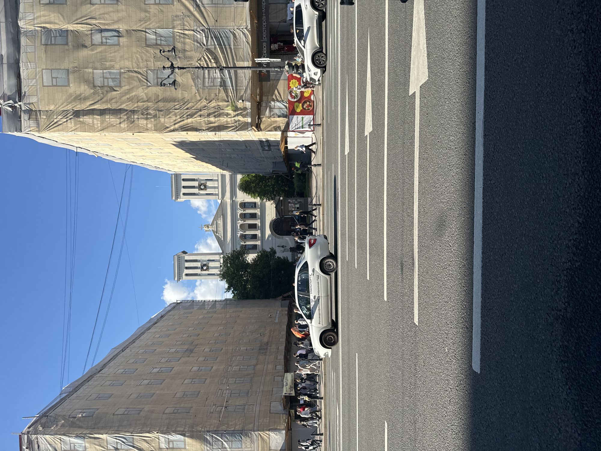 The image depicts a street scene in Saint Petersburg, Russia, likely on Nevsky Prospekt, a prominent avenue. The focal point is a white sedan with Russian license plates driving in the foreground, reflecting the clear blue sky. To the left and right of the street are large buildings, both covered in tan scaffolding and sheeting, indicating ongoing construction or renovation. A white church or cathedral with two tall bell towers and a classical facade stands in the background, partially obscured by the construction. A crowd of pedestrians is visible on the sidewalk in front of the buildings. The street itself is paved and marked with white lane lines and an arrow. The overall scene suggests a typical urban day with activity on the street and surrounding buildings undergoing refurbishment.