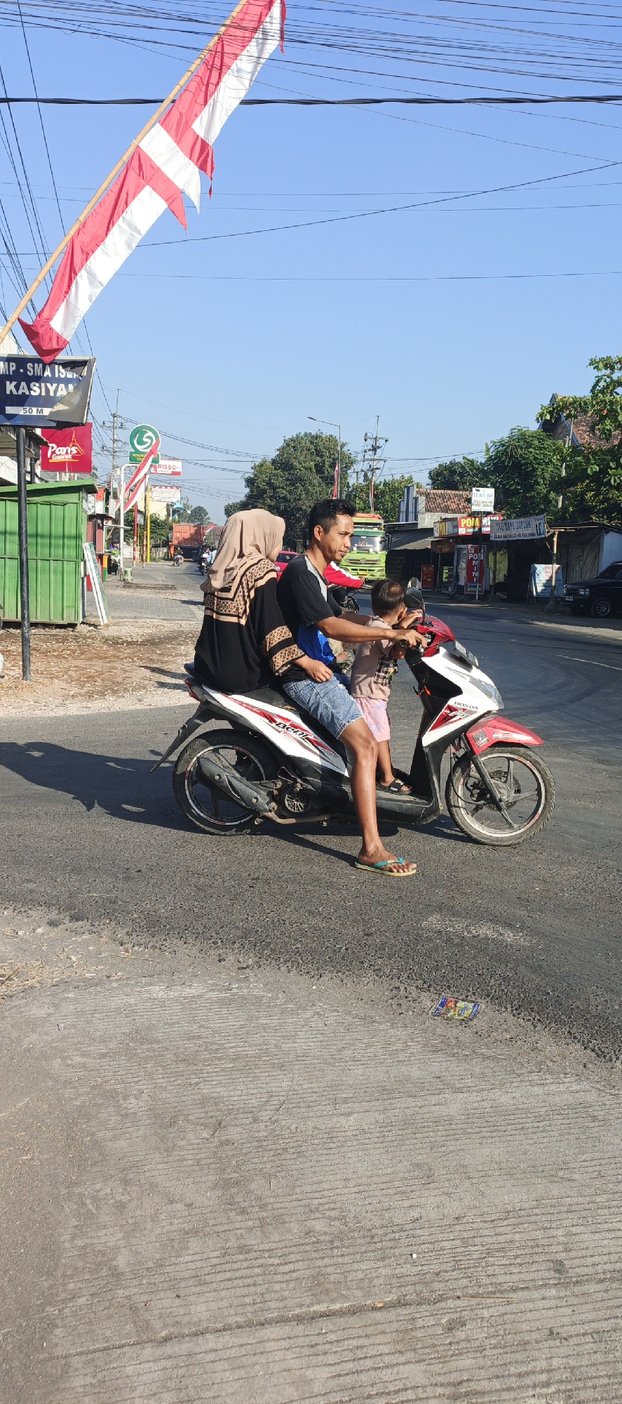 A family of three rides a red and white motorcycle down a paved road in Kasiyan, Indonesia. The father is driving, his son sits in front of him, and the mother is riding behind the father. The mother wears a beige hijab and a black and gold patterned dress. The father wears a black t-shirt and blue denim shorts. His son wears a striped shirt and pink shorts. The motorcycle is a Honda Beat. The road is lined with businesses and trees. A red and white Indonesian flag flies from a pole on the left side of the image. A large green garbage bin is visible on the left side of the road. Overhead power lines are strung across the sky. The sky is clear and blue. The lighting suggests it is a sunny day, likely late morning or early afternoon.