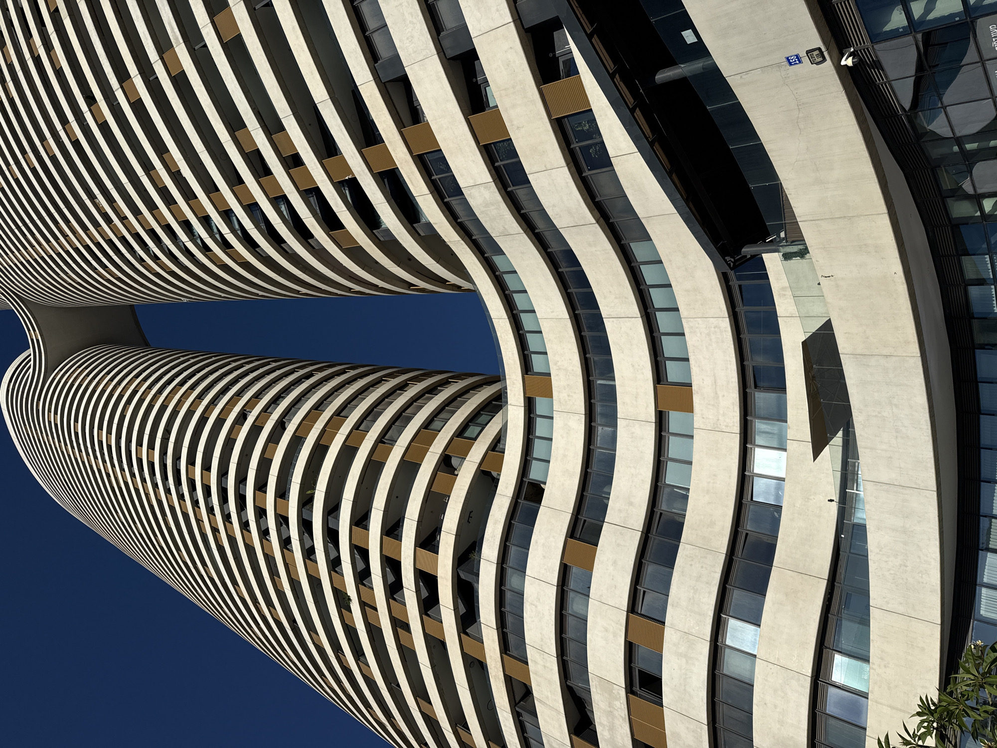 This is a low-angle, wide shot of the W Tower in Tel Aviv, Israel, on a clear, sunny day. The distinctive architectural design of the building features two curved, tall towers connected by a skybridge, with staggered balconies creating a wave-like facade. The exterior is a light-colored concrete, contrasted by horizontal bands of darker material and bronze-colored louvers on some windows. The sky is a deep, cloudless blue. The lower portion of the building shows multiple levels of windows and balconies, with some glass railings and a darker, covered area on the ground floor. A small green plant is visible in the bottom left corner. A street sign with the number "158" in blue and white is mounted on the building, along with a security camera. The building is a residential skyscraper and a hotel.