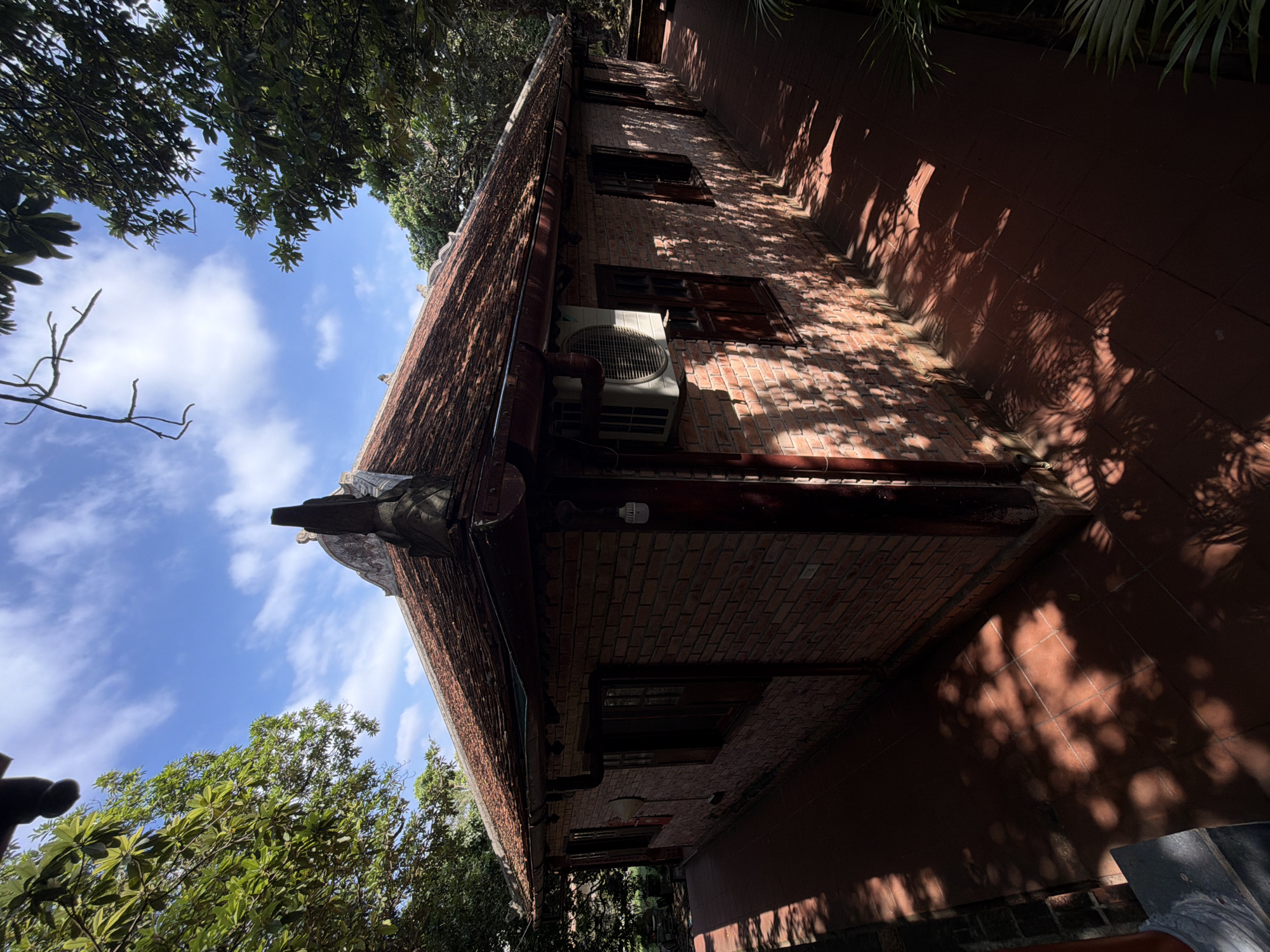 A low-angle shot shows a multi-story brick building in Thủy Xuân Ward, Vietnam. The building has a traditional tiled roof with a decorative gable. An air conditioning unit is mounted on the brick wall. The building casts shadows onto a terracotta-tiled ground. The sky is blue with scattered clouds, visible through a canopy of trees surrounding the building.
