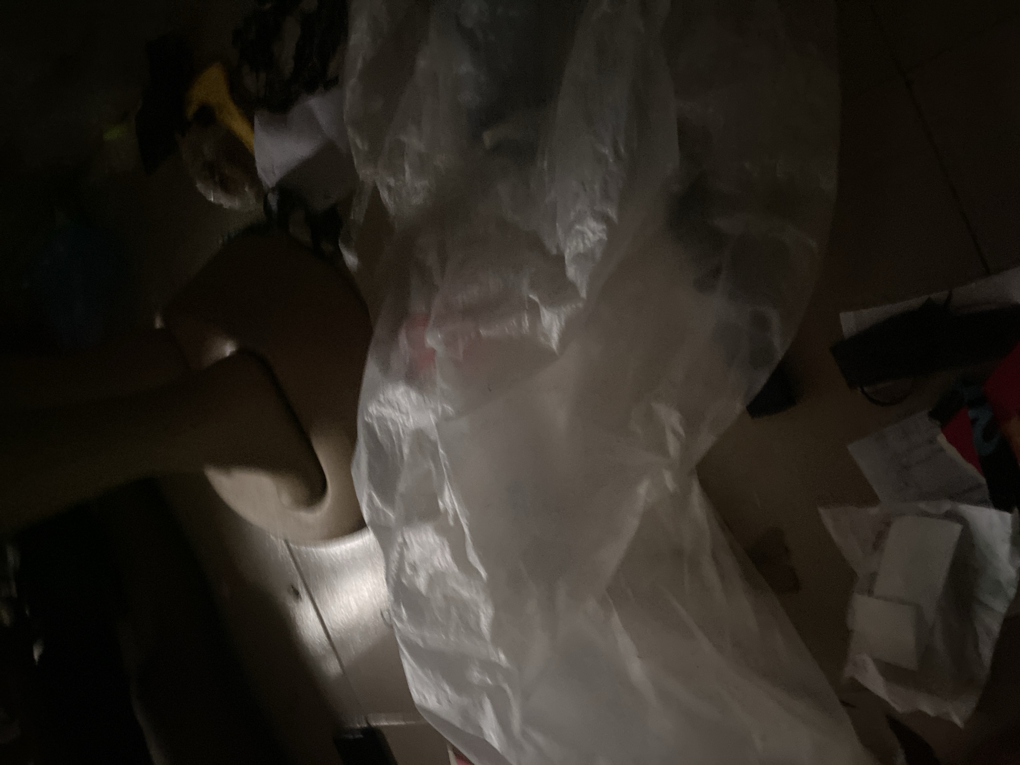 The image depicts a dimly lit scene on a wooden floor. In the foreground, a large, crumpled white plastic bag dominates the frame, partially obscuring a human leg bent at the knee. The leg appears to be resting on or near the plastic bag. The surrounding environment is dark and cluttered with various unidentifiable objects, some of which appear to be other pieces of plastic or fabric. Towards the bottom left of the image, a few white paper-like items are visible, possibly napkins or tissues, arranged in a loose stack. The lighting is uneven, with areas of deep shadow and some highlights reflecting off the plastic. The overall impression is one of disorder. No specific location in Jauro Yinu, Nigeria, can be determined from the visual information provided.