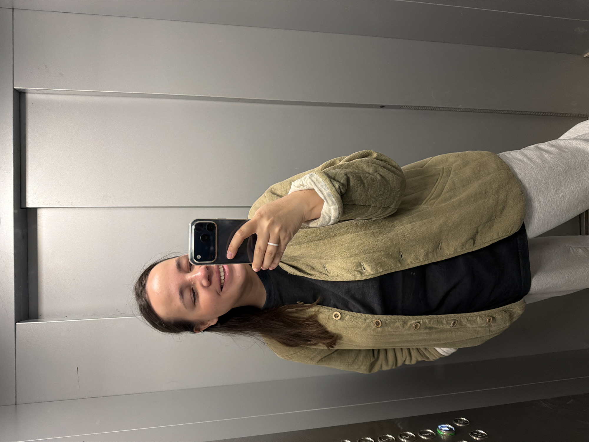 An image shows a young woman taking a selfie inside an elevator in Chaikovsky, Russia. The woman is smiling broadly with her eyes closed, reflecting a cheerful expression. She has dark hair and is seen from the chest up, although her lower body wearing light-colored pants is also partially visible. She is dressed in a dark t-shirt layered under a light olive-green, quilted jacket with decorative buttons. A simple white ring is visible on her left ring finger.
She holds a black smartphone with multiple camera lenses in her right hand, which is visible in the reflection as she captures the image. The elevator interior features sleek, metallic-gray walls with horizontal paneling and vertical seams, consistent with a modern lift. At the bottom right of the frame, a portion of the elevator's control panel is visible, displaying buttons for different floors (e.g., "1", "2", "3"), with one button, possibly "4", illuminated with a green-blue light. The lighting appears bright and even within the confined space.