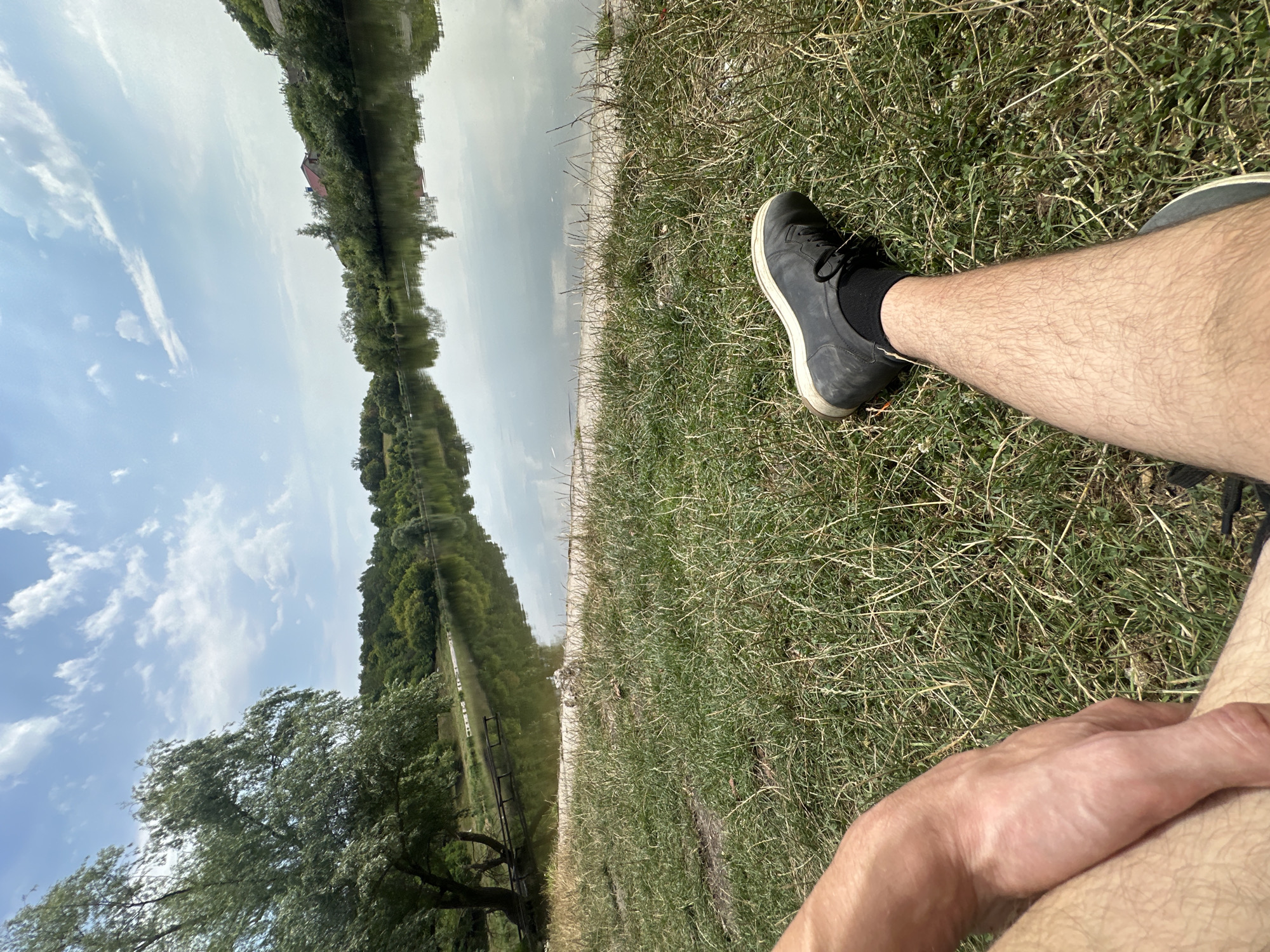 This is a first-person perspective, eye-level shot of a person sitting on the grass near a lake. The person is wearing a black shoe and black sock on their right leg, which is extended forward. A portion of the person's left arm and thigh are also visible. The background shows a calm lake reflecting the sky and surrounding trees. On the far bank of the lake, there is a dense line of trees and a few houses. The sky is partly cloudy with white clouds scattered across a blue expanse. The environment appears to be a natural setting, possibly a park or recreational area in or near Makiv, Ukraine.