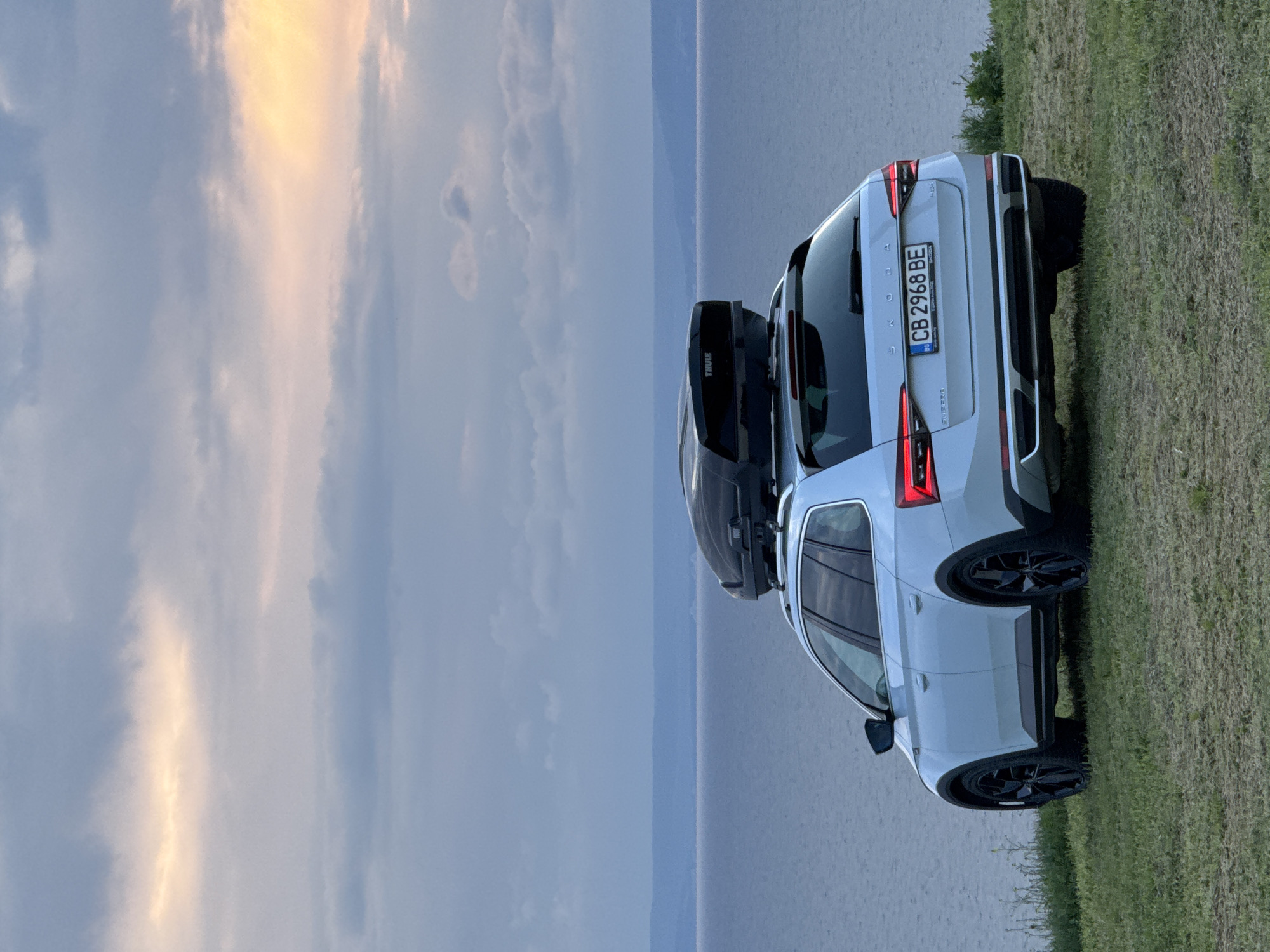A white Skoda Superb, with a Thule roof box, is parked on a grassy area overlooking a body of water, potentially the Black Sea, with a view of the horizon under a partly cloudy sky, possibly near sunset. The car's license plate, "CB 2968 BE," is visible. The location is presumed to be near Balgarevo, Bulgaria, based on the request.