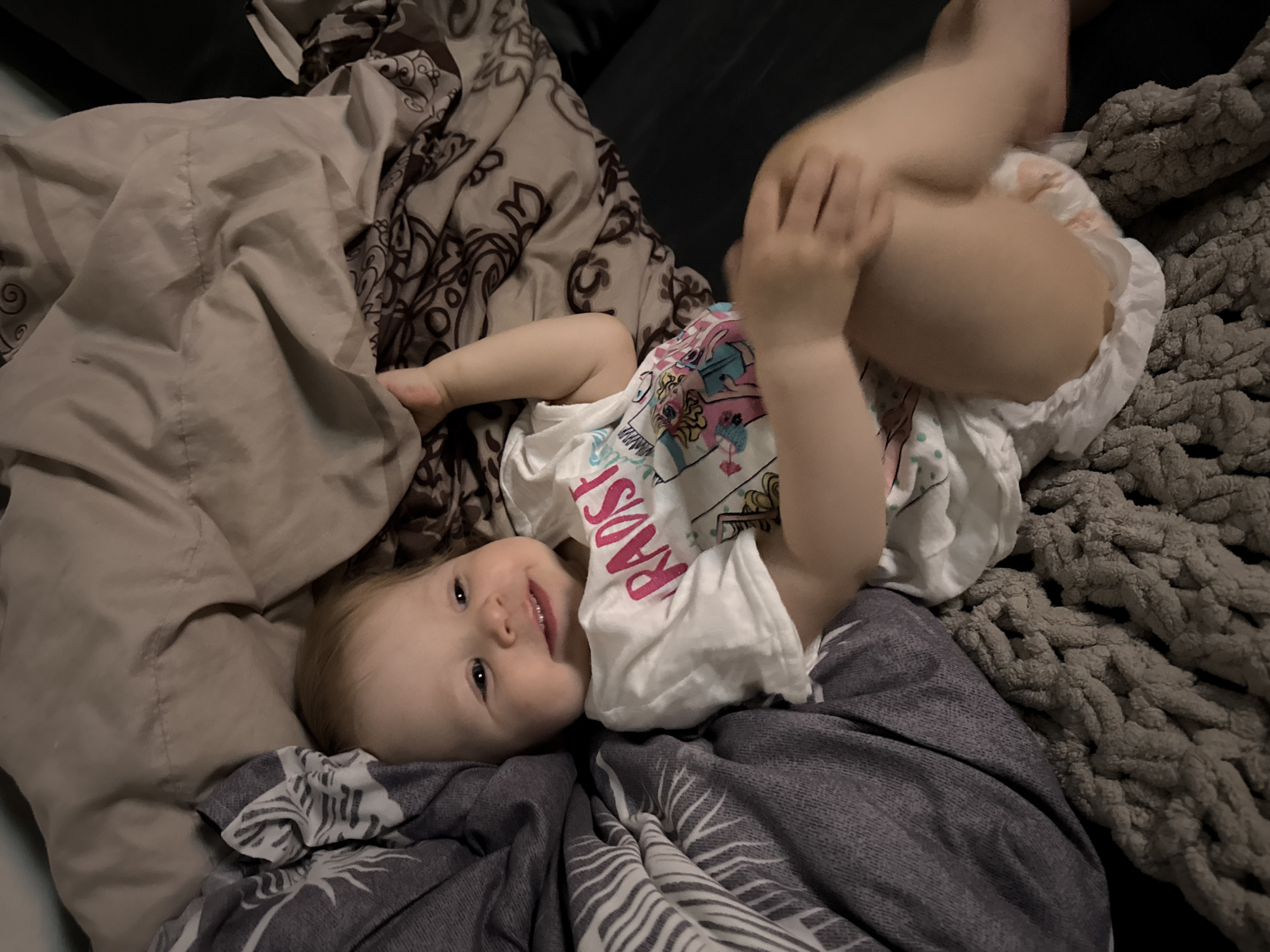 A baby wearing a white t-shirt and a diaper is lying on a bed covered with a grey and white striped blanket, a beige blanket, and a gray braided blanket. The baby is holding their legs up in the air. The location is assumed to be a private residence in the city of Chaikovsky, Russia.