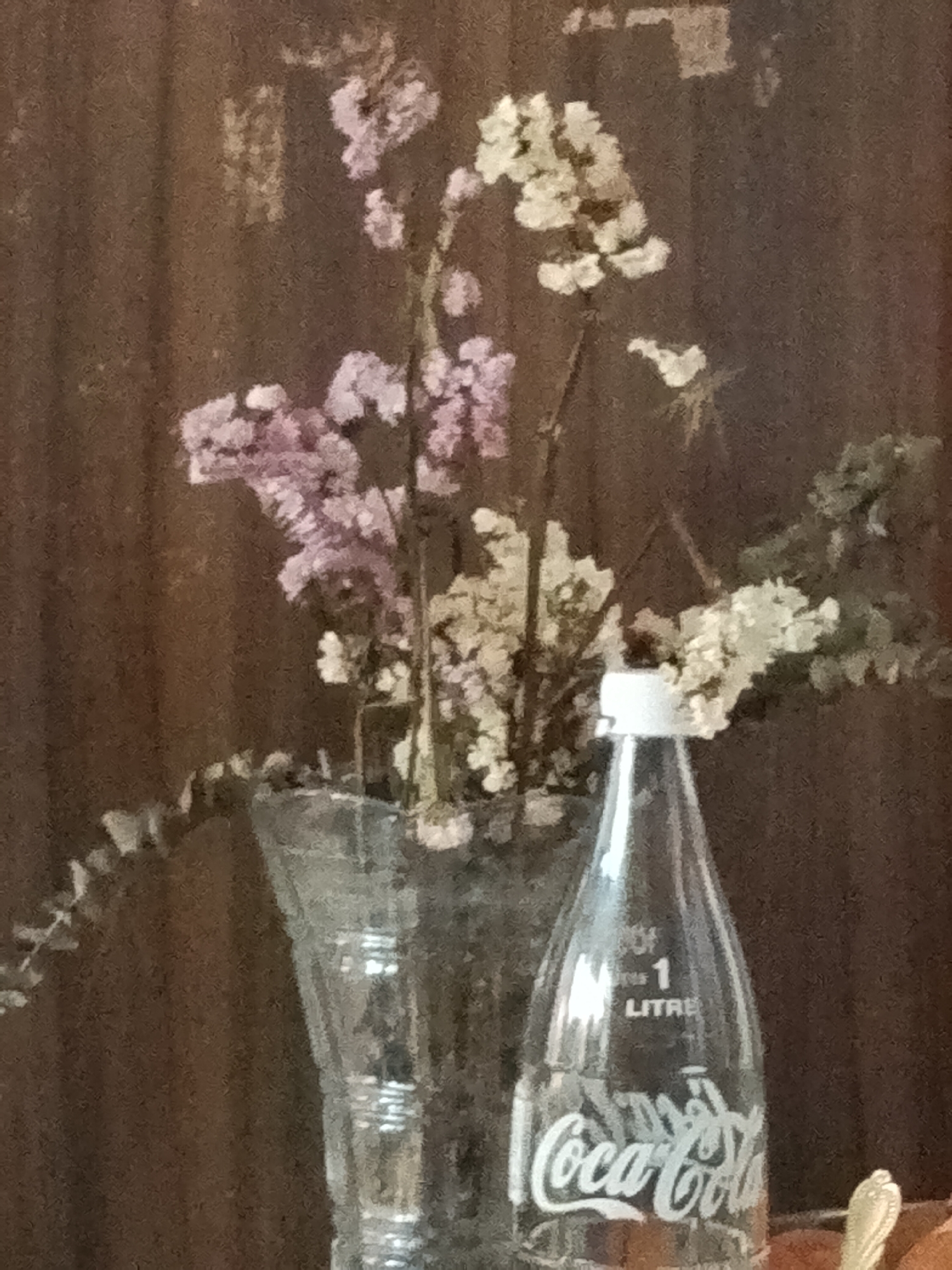 The image displays a still life featuring a bouquet of dried flowers and an empty glass Coca-Cola bottle, set against a dark, textured background that appears to be wooden paneling.
On the left, a clear glass vase holds a collection of dried flowers. These include small blooms in muted shades of light purple/pink and white/cream, intertwined with some dried green foliage, suggesting varieties like statice or similar. The flowers appear brittle and preserved rather than fresh.
To the right of the vase is an empty, clear glass Coca-Cola bottle with a white plastic cap. The iconic white "Coca-Cola" logo in its distinctive script is prominently featured on the lower body of the bottle. Molded into the glass above the logo, the text "OF 1 LITRE" is clearly visible, indicating its volume.
The background is a uniform dark brown, likely wood grain, which is slightly out of focus, providing a soft backdrop to the main subjects. In the bottom right corner, a small, ornate handle of a piece of cutlery, possibly a spoon, is partially visible. The overall lighting is soft and indirect, creating a subdued and somewhat rustic atmosphere. The scene suggests an indoor setting.