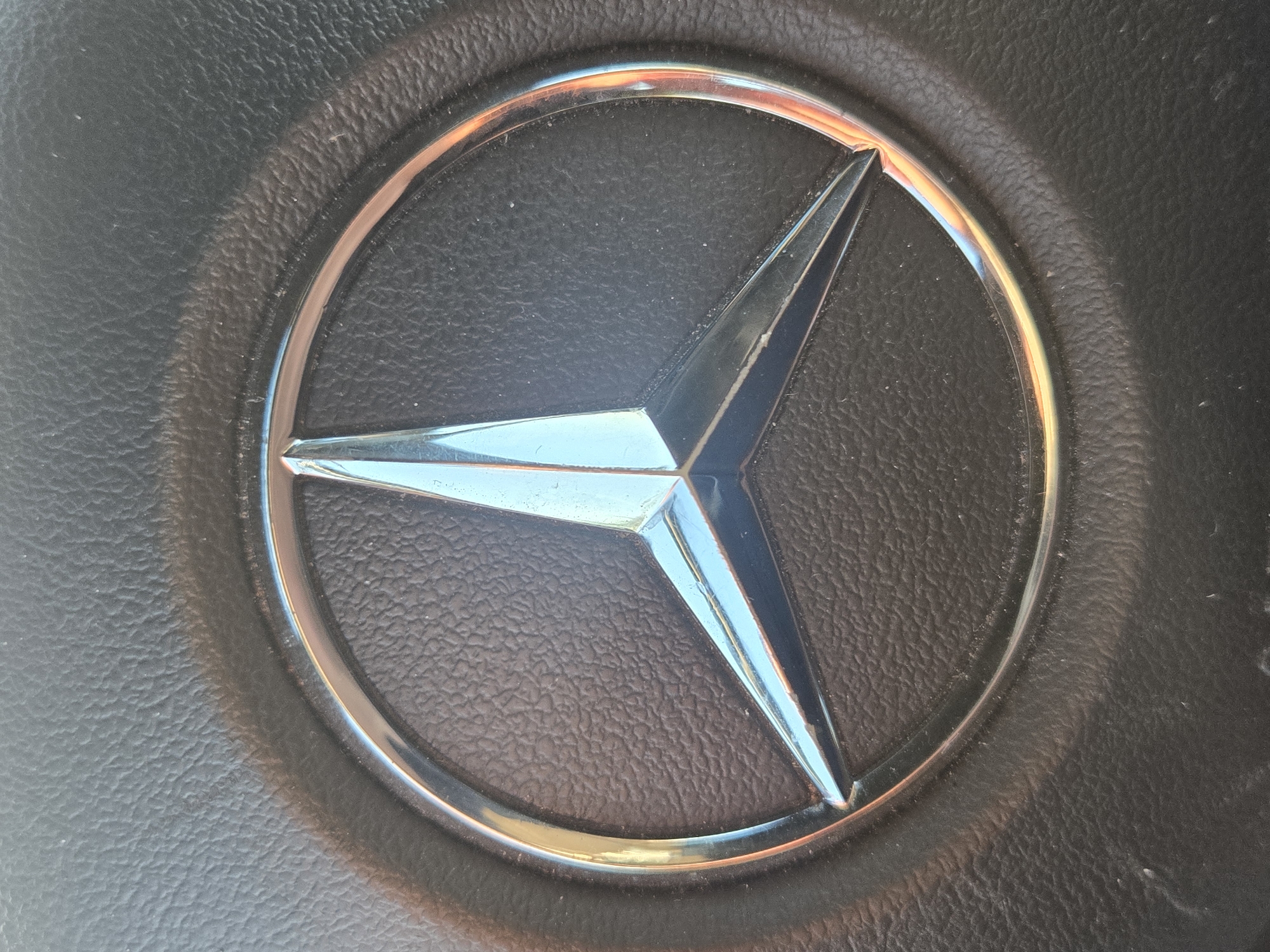 A close-up of a Mercedes-Benz logo centered on a dark, textured surface, likely a steering wheel. The logo is a silver circle encompassing a three-pointed star. The environment is indeterminate. The location is unknown.
