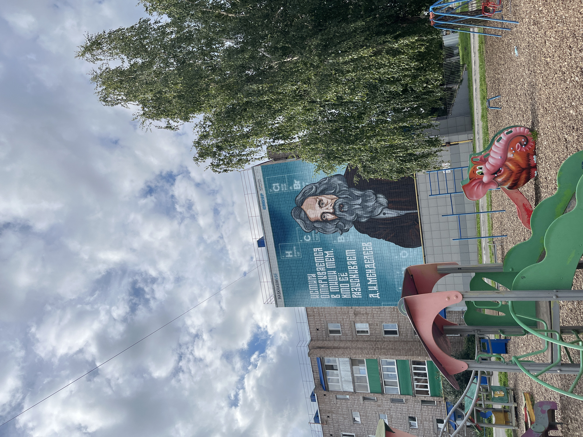 The image shows a mural of Dmitri Mendeleev on the side of a building in Blagoveshchensk, Russia. The mural is on a large, light blue wall that is part of a modern building with several windows. The mural depicts Mendeleev with a long beard and gray hair, wearing a dark jacket. Text in Russian is visible on the wall, featuring chemical elements like Hydrogen (H) and Carbon (C). To the left of the mural is a multi-story residential building with light-colored brickwork and some green and white accents on the lower floors. In the foreground, there is a playground with colorful slides, swings, and a whimsical elephant-shaped play structure. A large tree with green foliage is positioned to the right of the mural, partially obscuring the building. The sky above is partly cloudy.