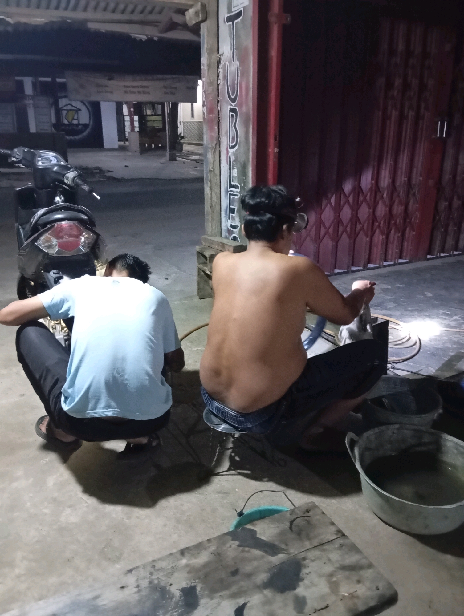 Two individuals are crouched on a concrete surface in what appears to be an outdoor or semi-enclosed area, possibly a shop front or workshop, in Leman Benawi, Indonesia. The lighting suggests it is nighttime.
The person on the left is wearing a light blue t-shirt and dark pants, with their back to the camera. They are looking down towards something in front of them. They are wearing dark sandals.
The person on the right is shirtless, wearing dark patterned shorts. They are facing away from the camera and appear to be holding an object in their hands, possibly engaging in a task that involves a bright light source emanating from the area in front of them. They are wearing eyeglasses on their forehead.
In the background, a motorcycle is parked to the left. To the right, there is a large red metal rolling door. Various objects are scattered on the ground, including buckets, one of which contains water, and a wooden board. A banner is visible on a building in the background. The overall environment suggests a utilitarian or working space.