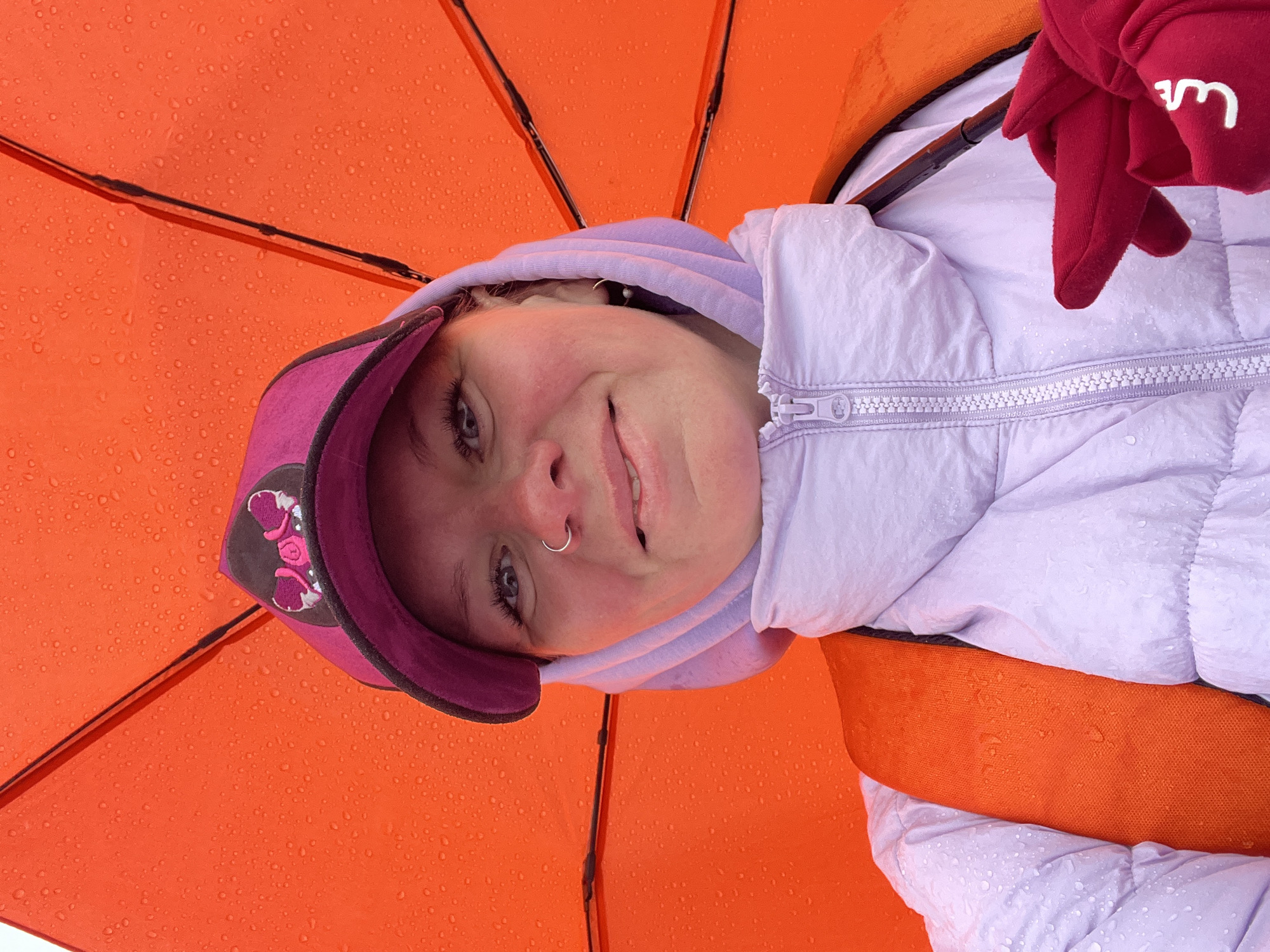 The image is a low-angle shot, looking up at a young person smiling directly at the camera while standing under a bright orange umbrella. The umbrella's canopy is visibly covered in numerous water droplets, indicating rain. The person wears a light purple puffer jacket over a lavender hoodie, and a maroon baseball cap with an embroidered pink and black design. They have a silver nose ring on their left nostril, and their eyes are blue. Their cheeks appear rosy, and they have a joyful expression. On the right side of the frame, a hand in a red glove or mitt is visible, holding a part of the umbrella structure. Inverted white text, possibly reading "wM" or similar, is present on the red fabric of the glove. The scene suggests an outdoor setting during a rainy day in Chaikovsky, Russia, with the person appearing happy despite the weather.