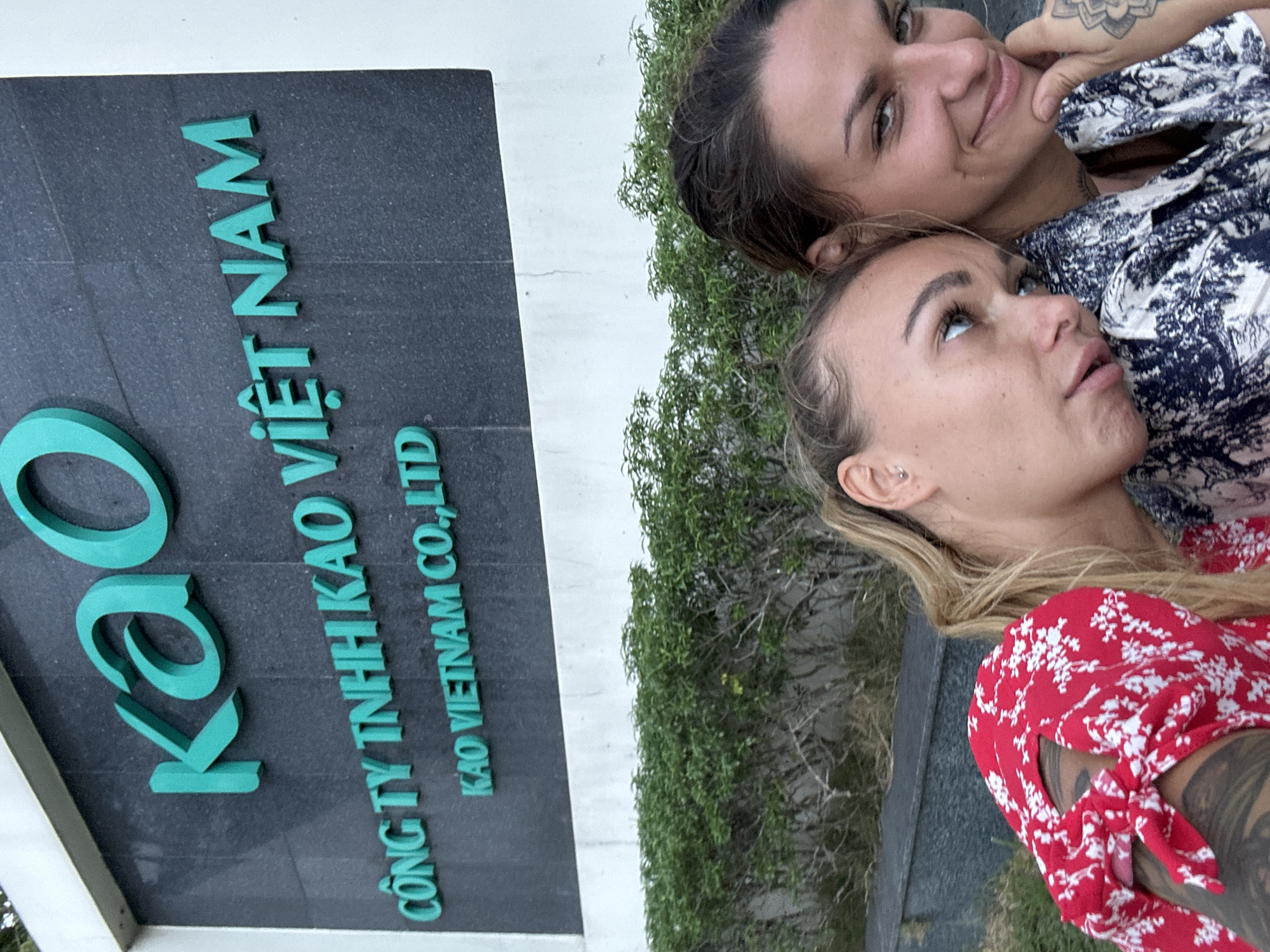 Two women are positioned beside a sign that reads "Kao," "Công ty TNHH Kao Việt Nam," and "Kao Vietnam Co., Ltd." A green shrubbery is situated between the sign and the women. The location is likely in Đồng Nai, Vietnam, given the Vietnamese text on the sign.